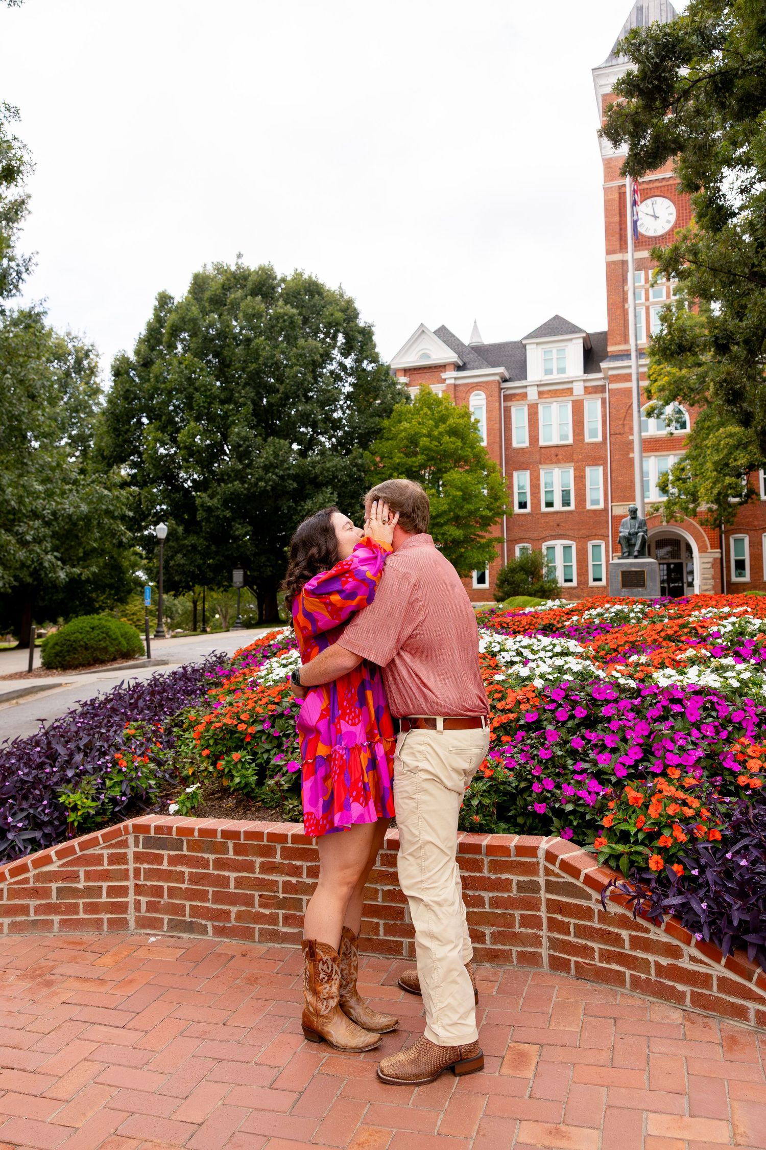 Couple kissing in front of colorful flower garden near brick clock tower building on university campus.