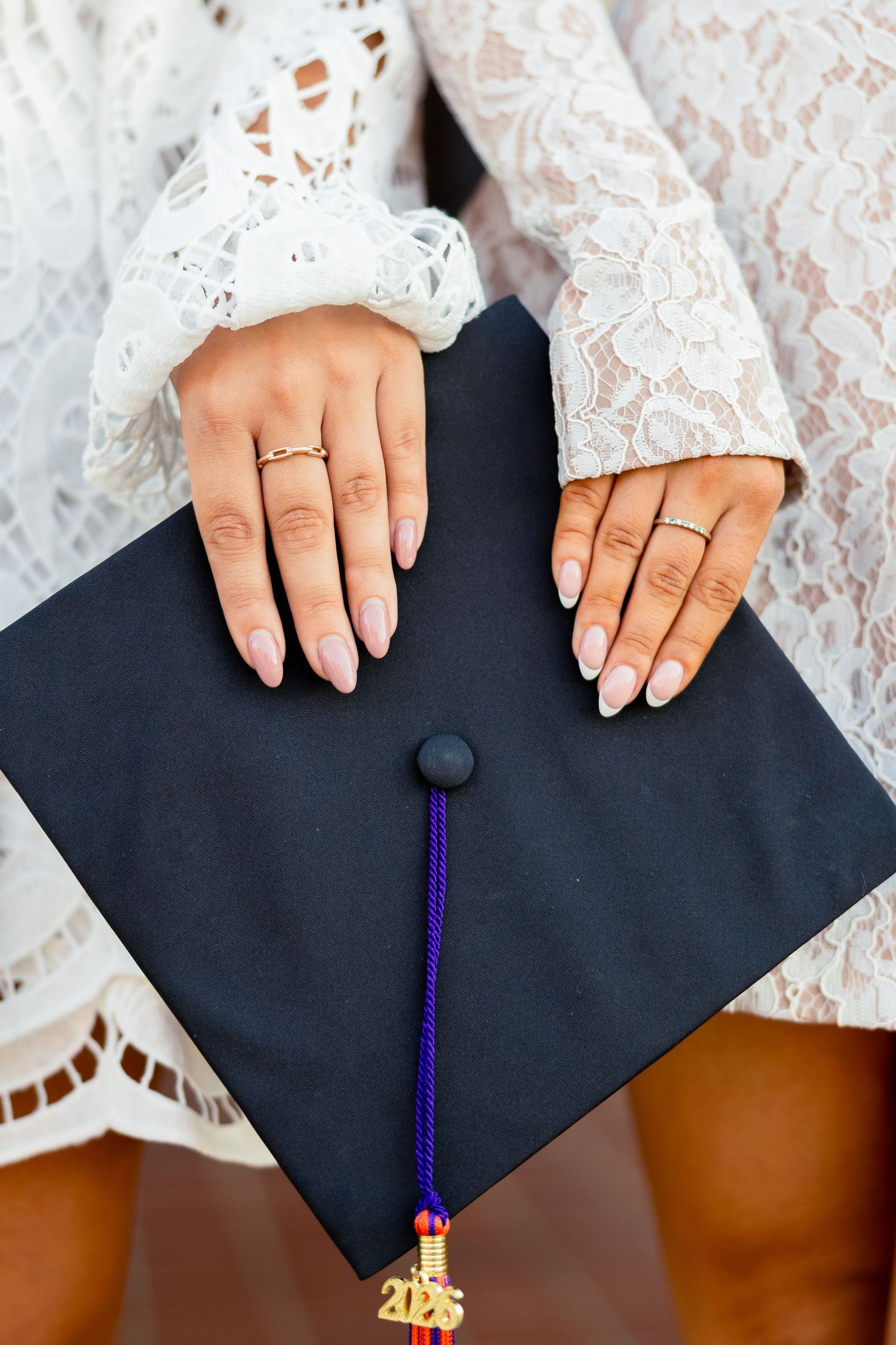 Close-up of hands with gold rings holding a black graduation cap with blue and gold tassel, wearing white lace dress.