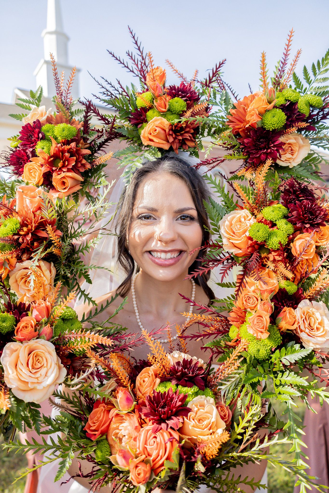 Smiling bride framed by a large autumn floral wreath with orange roses, burgundy blooms, and green foliage outdoors.
