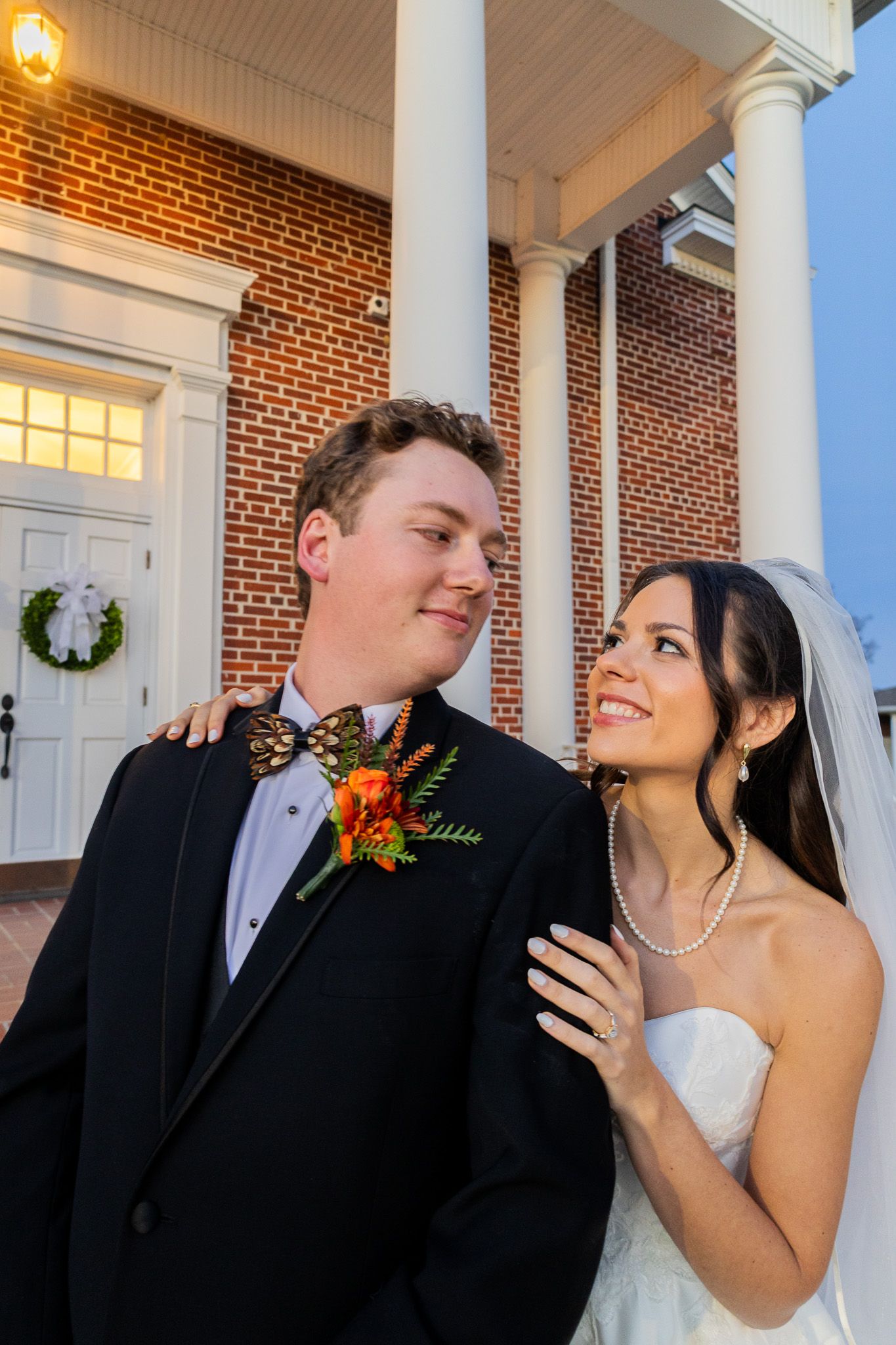 Bride in white gown and veil smiles at groom in black tuxedo with floral boutonniere outside brick wedding venue.