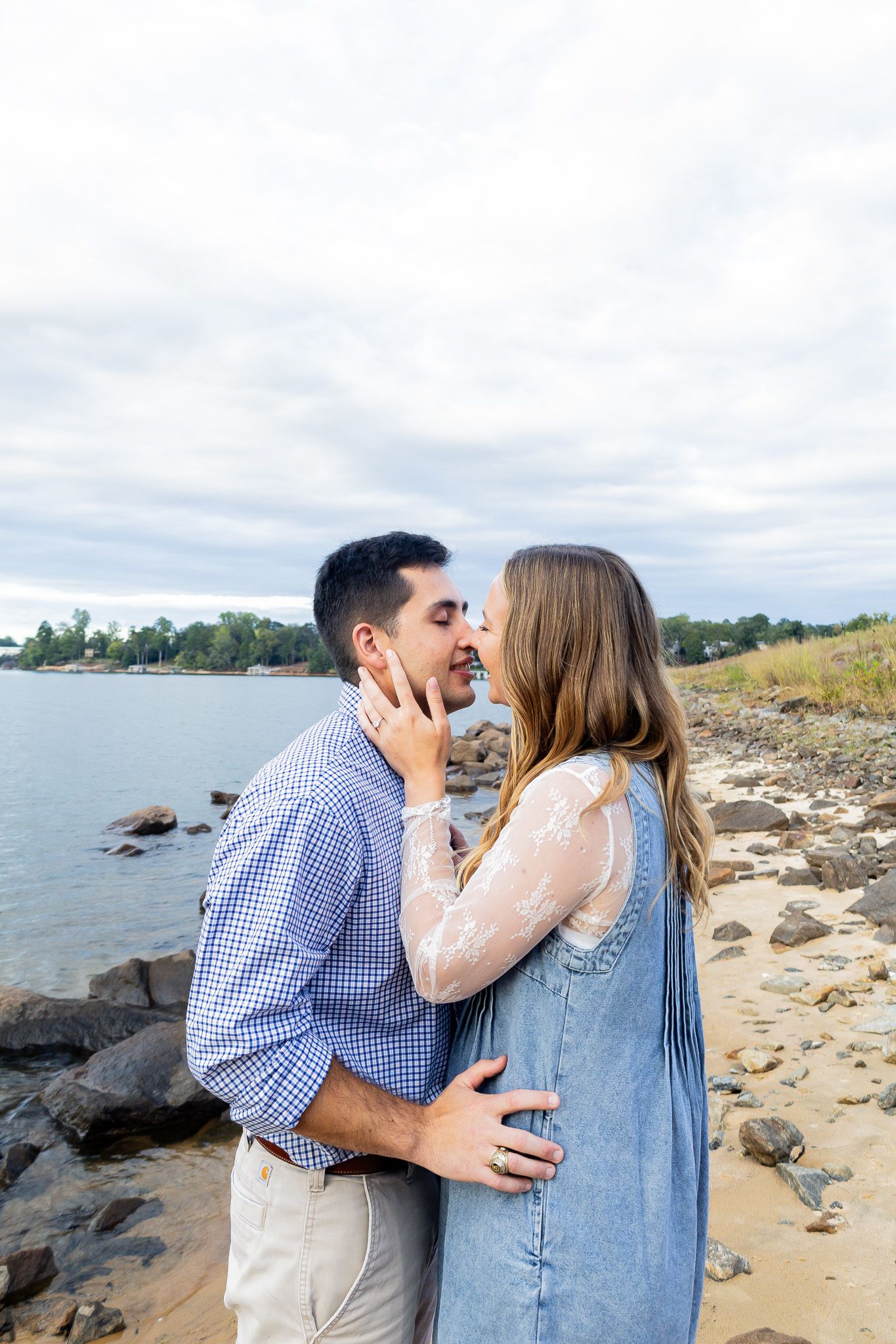 A couple shares a romantic kiss on a rocky beach shoreline, with the woman wearing a denim dress and lace top.