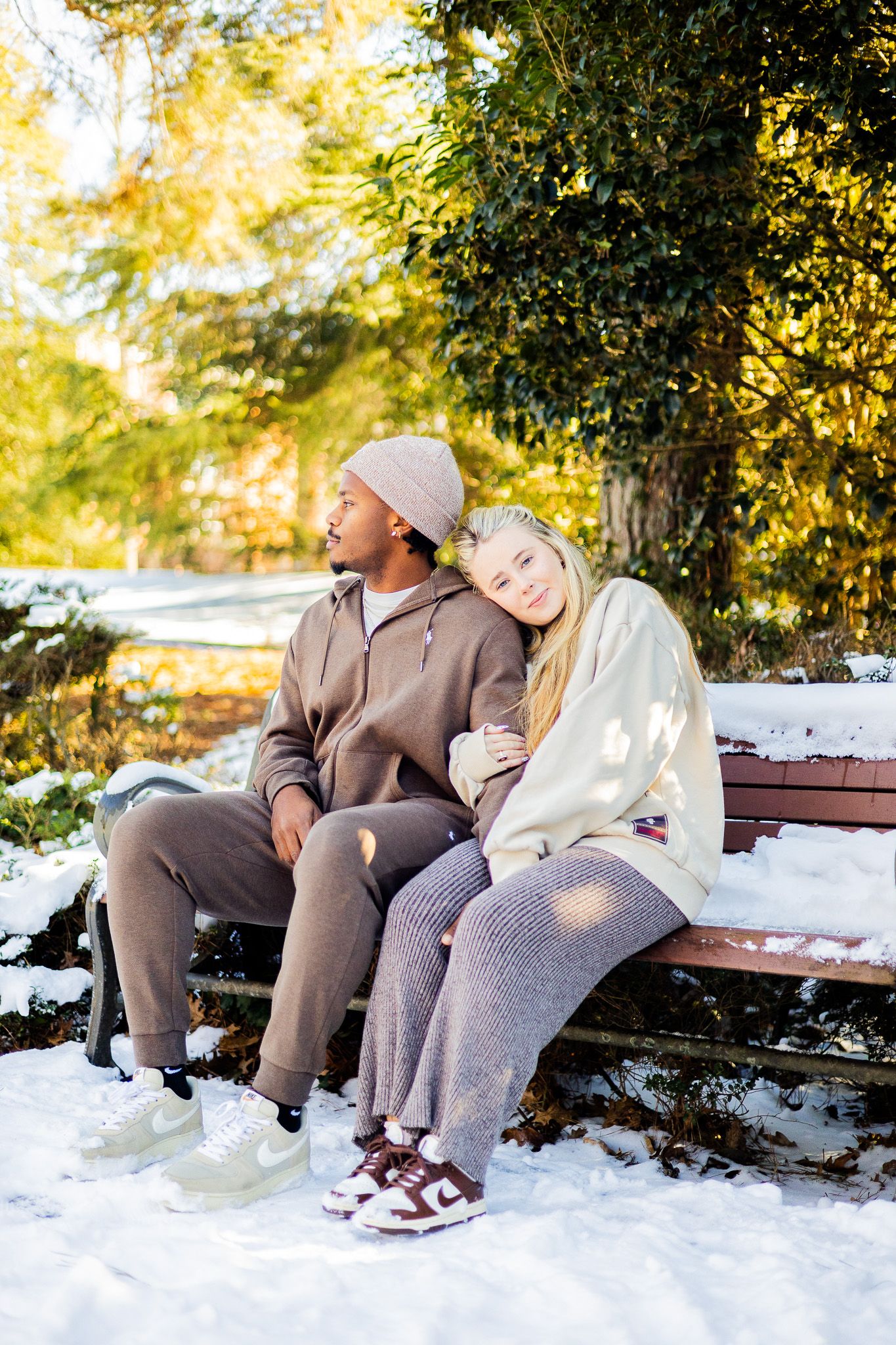 Couple sitting on a snowy park bench in winter, woman leaning on man's shoulder, surrounded by autumn trees.