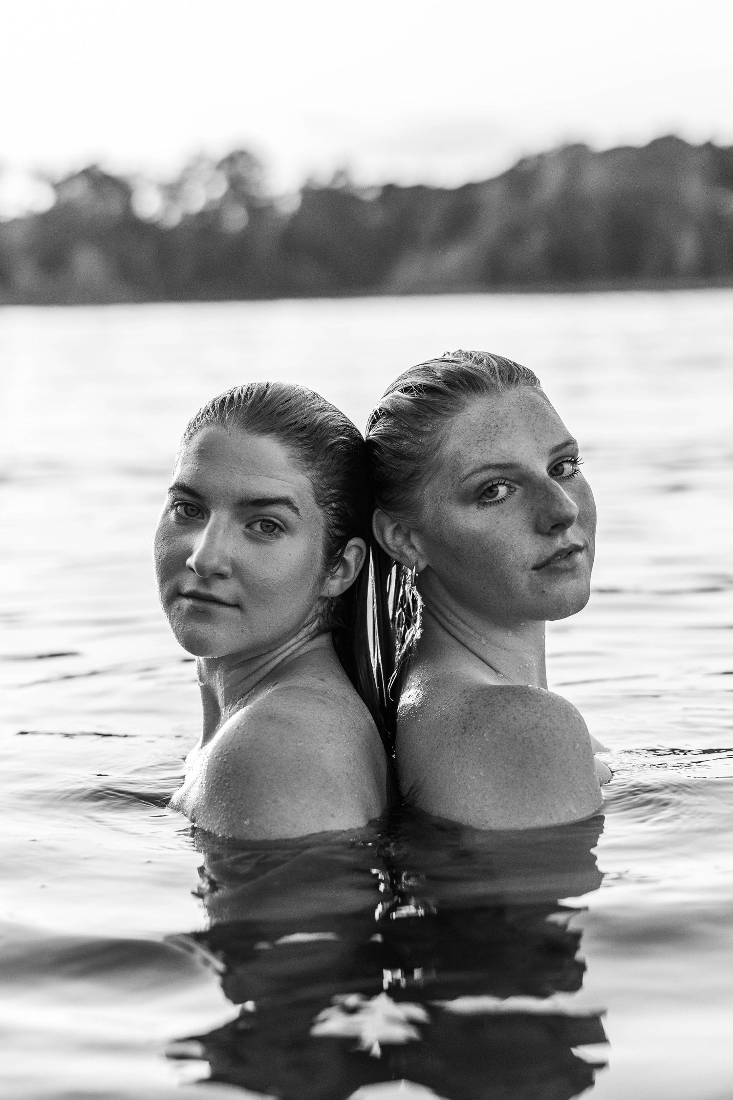 Two women standing back-to-back in a lake, wet hair, serious expressions, black and white portrait photography.