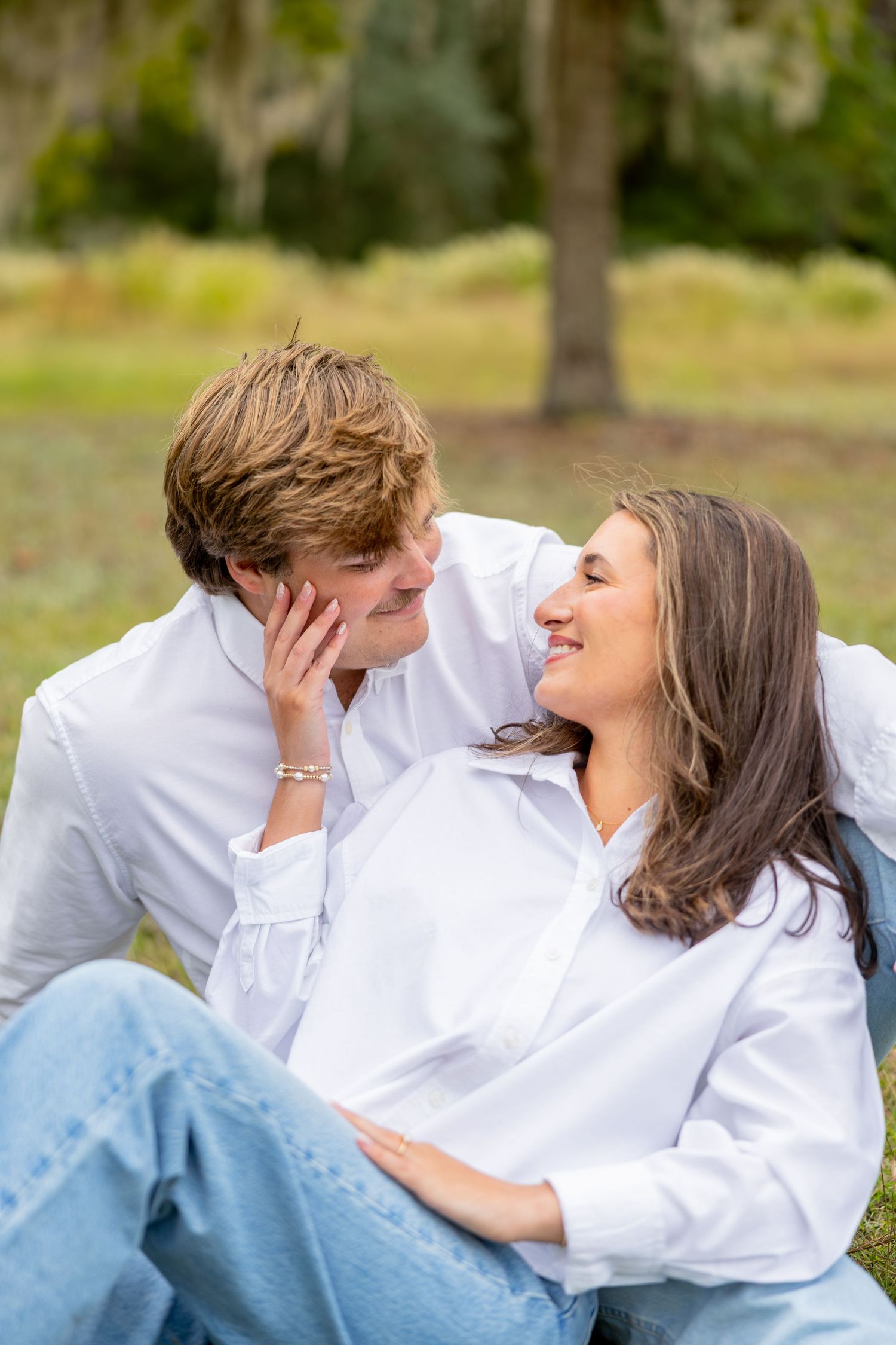 Smiling couple in white button-up shirts and jeans sit close together on a grassy lawn during an outdoor portrait session with trees behind.