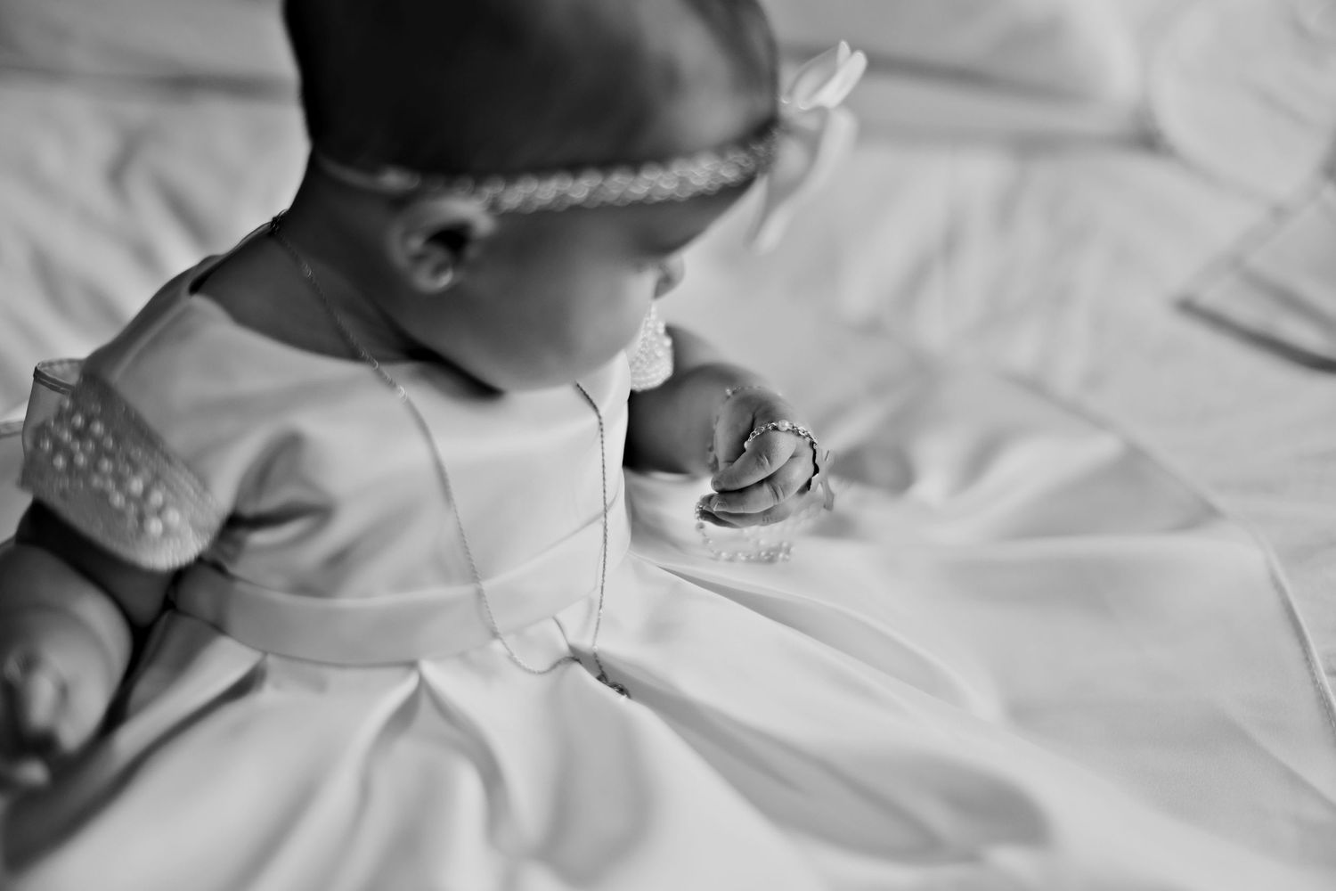 Black and white photograph of a small child in a white dress admiring a necklace while sitting on white fabric.