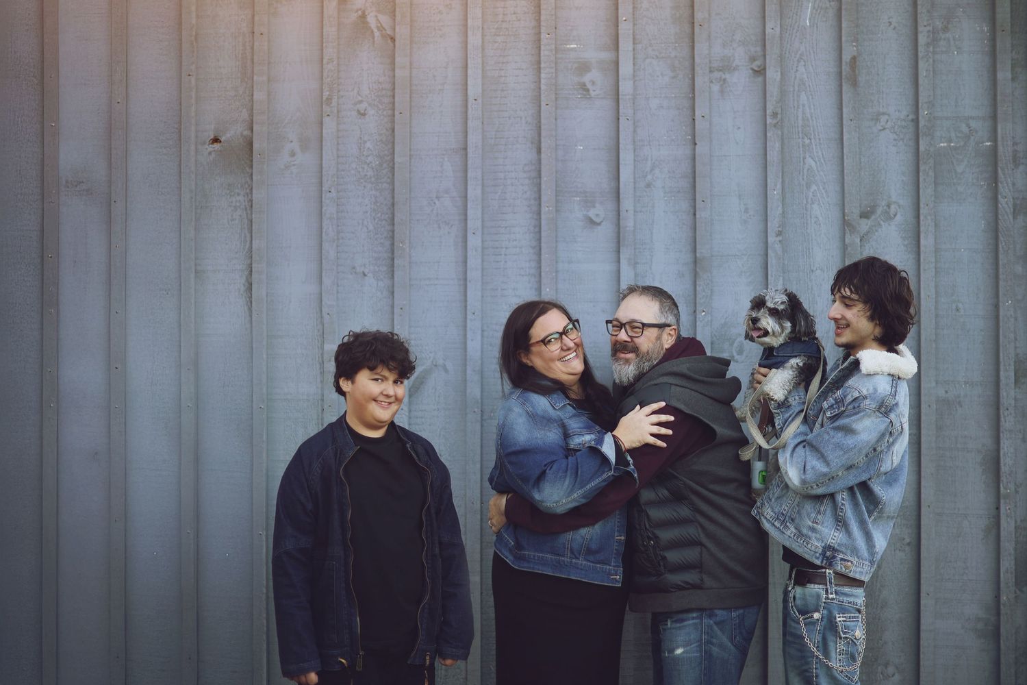 A group of friends in casual attire stand together and embrace against a corrugated metal wall backdrop.