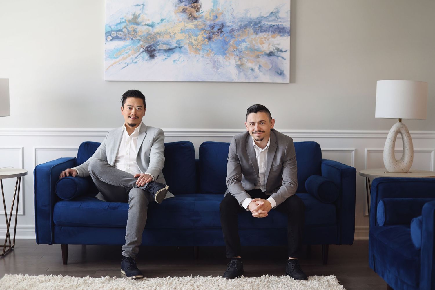 Two realtors sit on a dark blue sofa beneath an abstract artwork in a modern office setting.