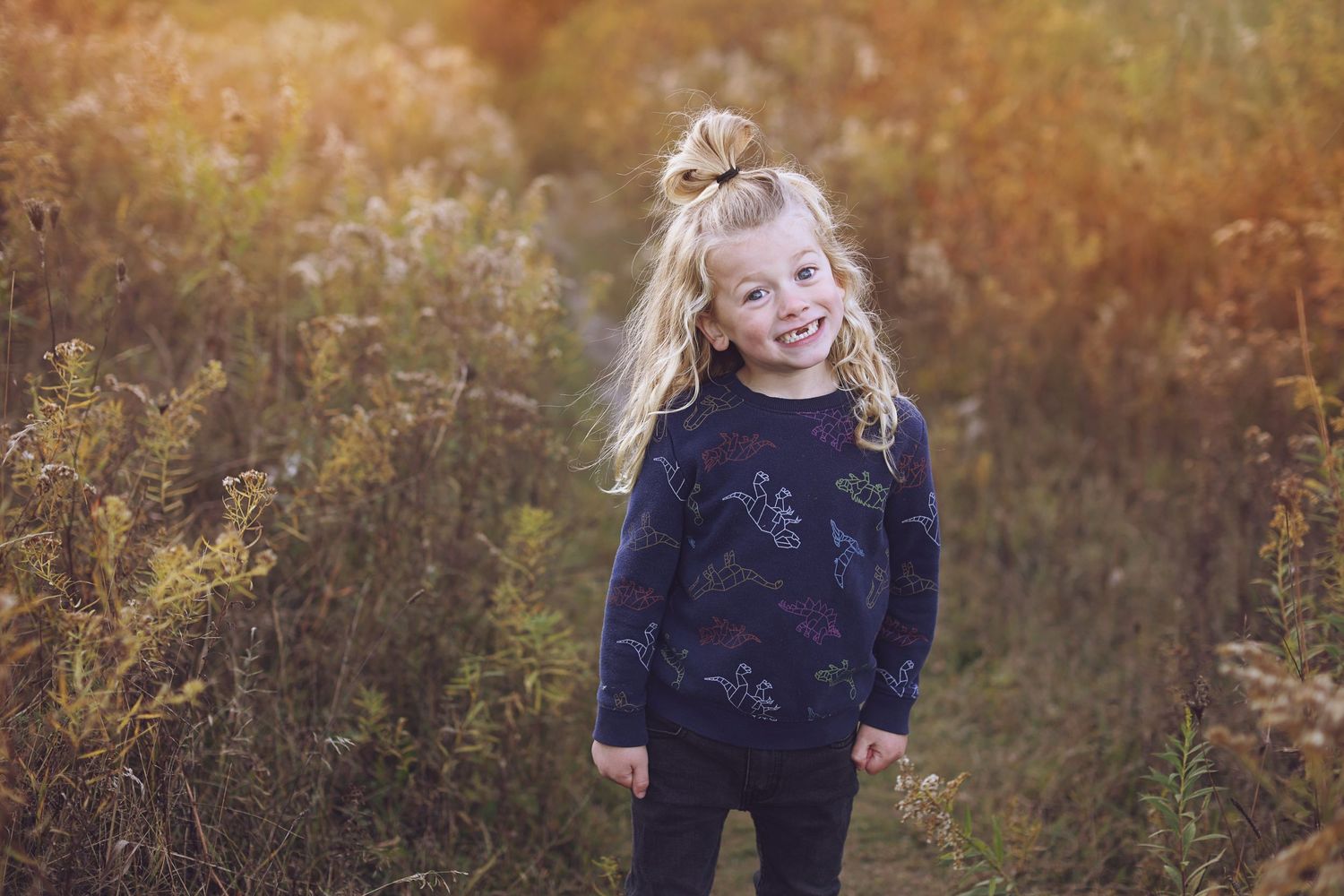 Child in dark sweater stands in golden autumn field with wild flowers during sunset.