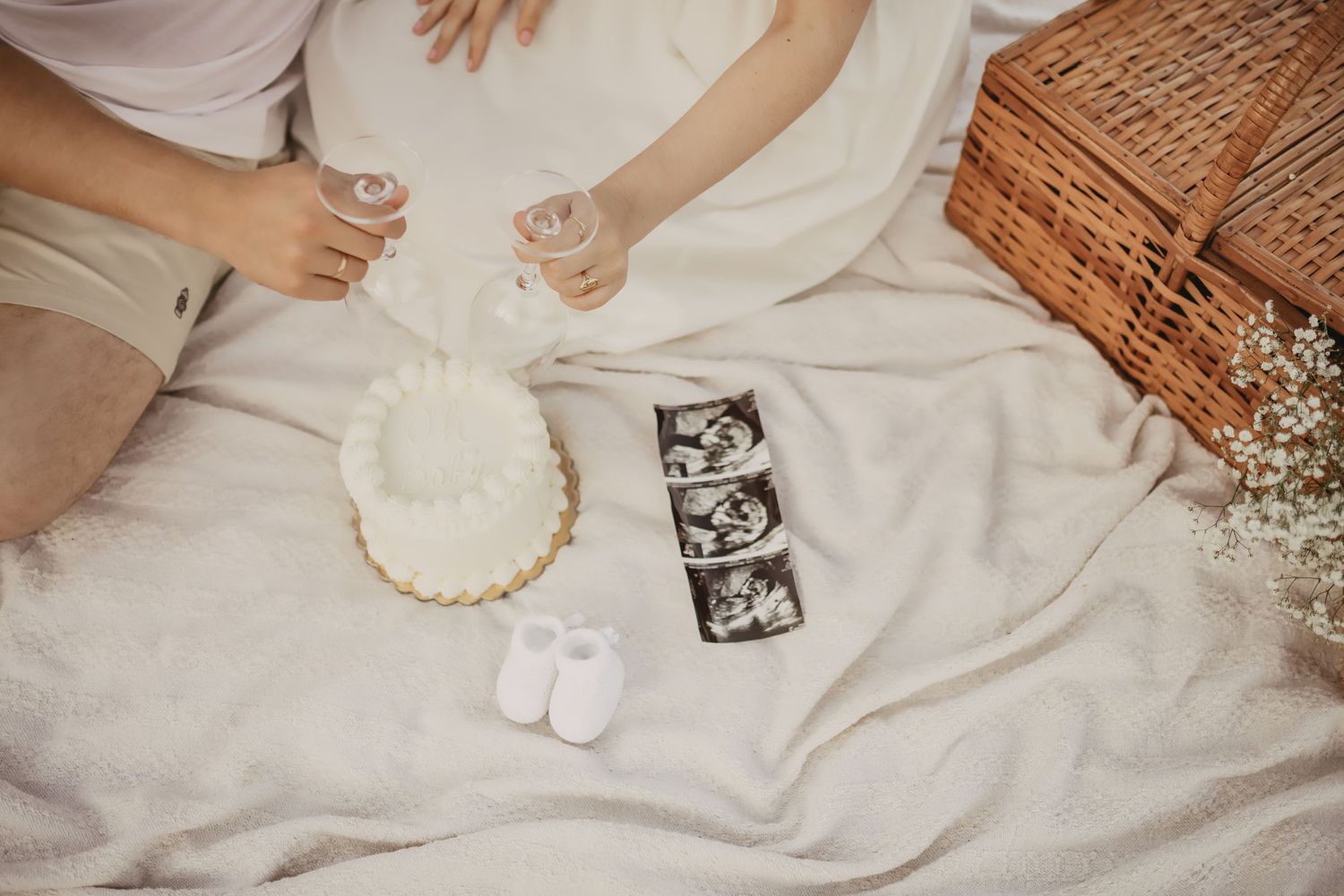 Baby announcement scene with ultrasound photos, tiny white booties, and floral crown laid on neutral fabric beside wicker basket.