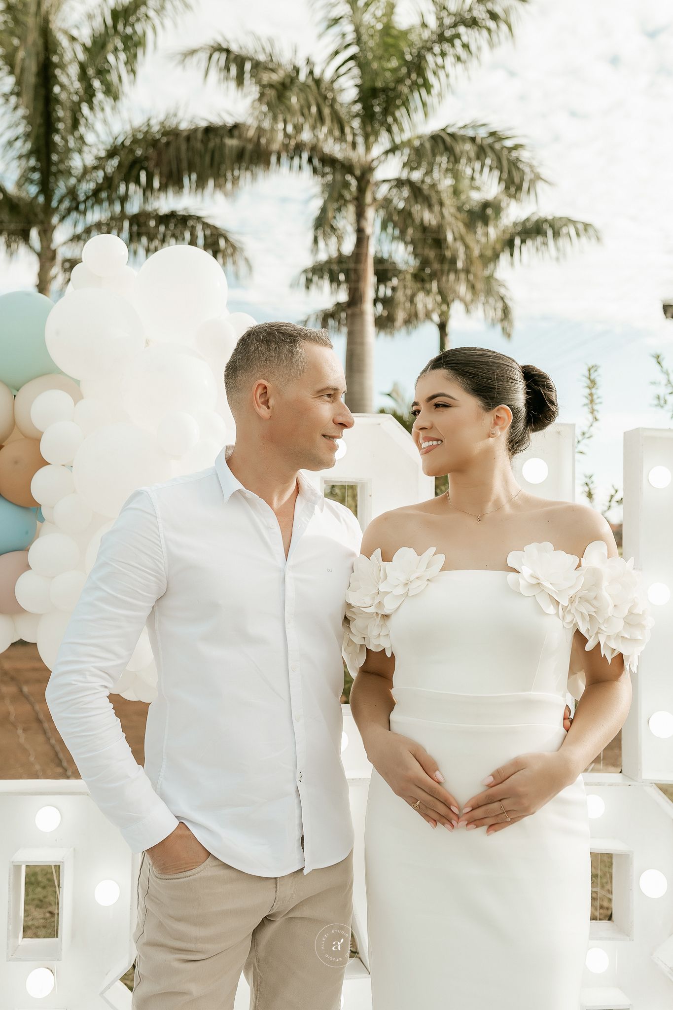 A romantic beach wedding scene with a couple in white attire embracing under palm trees and white balloons.