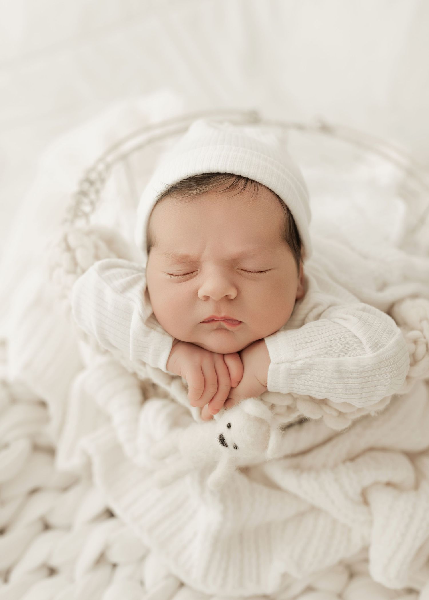 A sleeping newborn baby wearing a white knit cap rests peacefully on soft white blankets and textiles.
