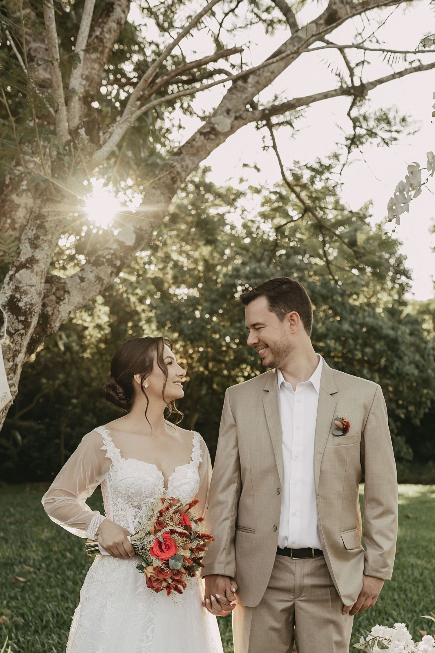 A couple in wedding attire shares intimate moments outdoors with sunlight filtering through trees in the background.