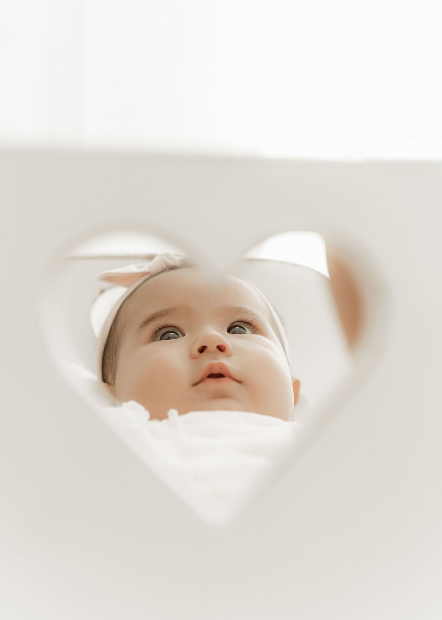 A baby wearing a white cap gazes upward through a heart-shaped cutout in a white background.