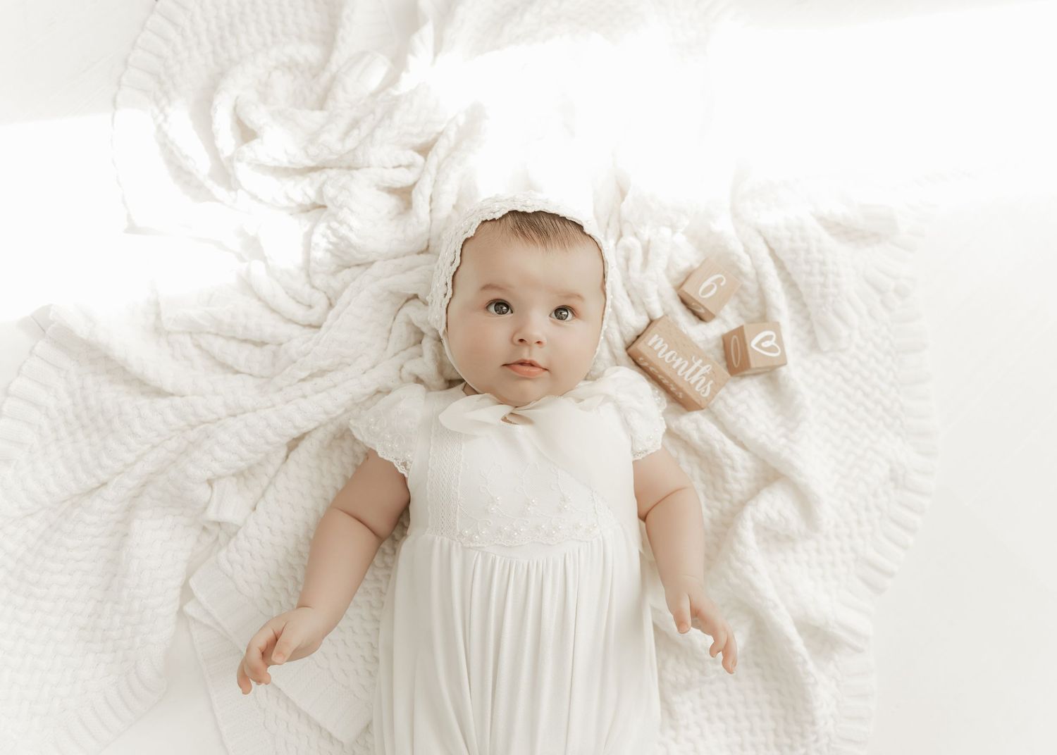 Baby wearing white dress lying on white textured blanket with plush teddy bear toy.
