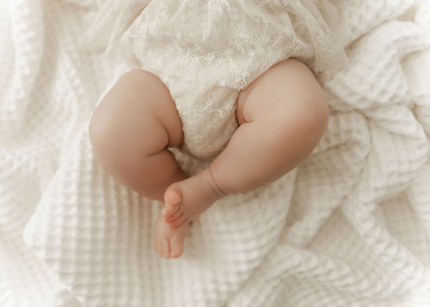 Tiny baby feet and legs resting on soft white textured blankets in gentle lighting.