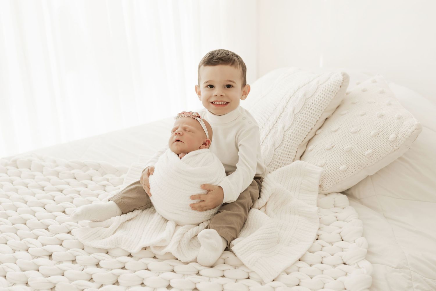 Two siblings in white clothing sitting on a chunky knit blanket with white pillows in a bright studio setting.