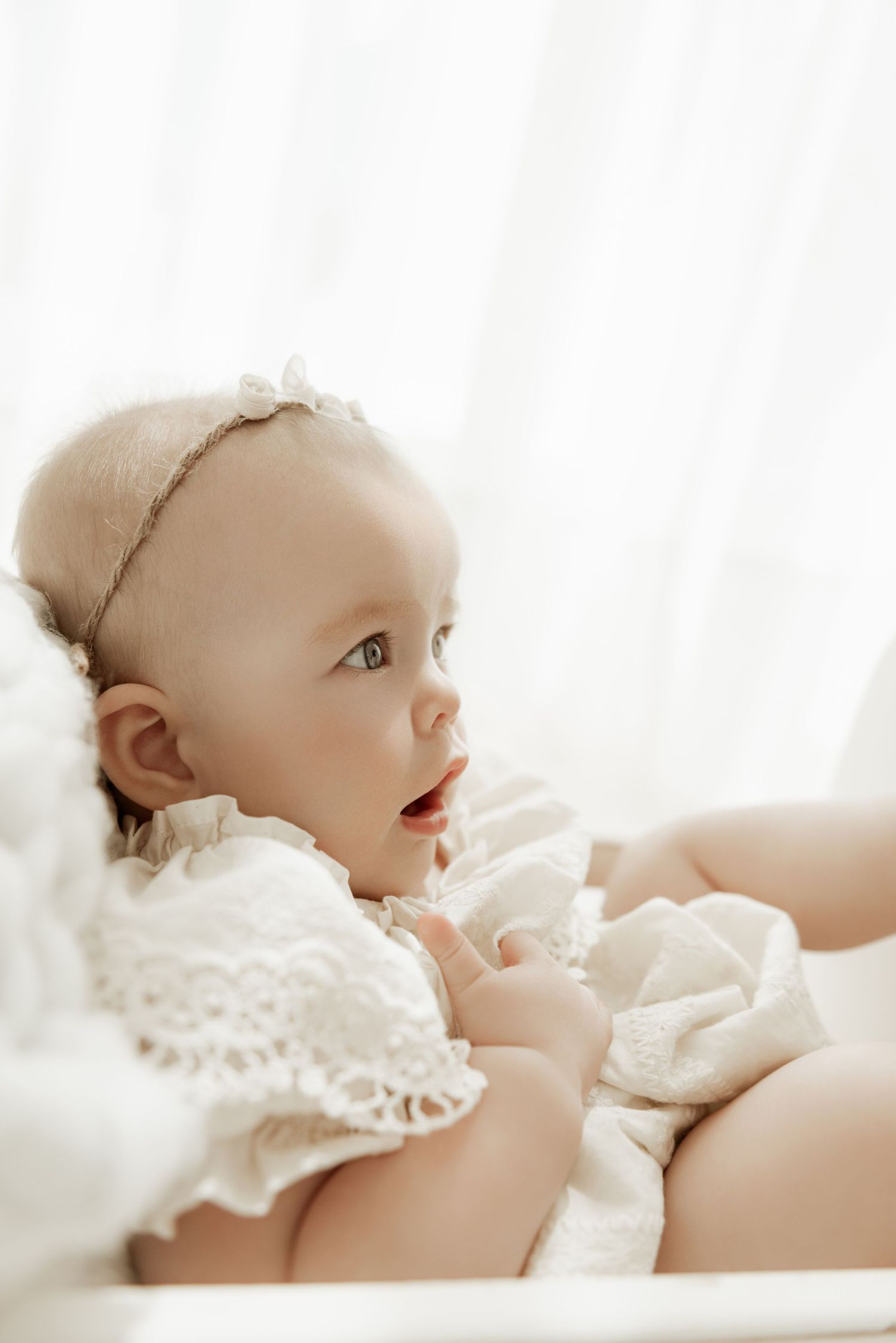 A baby in white lace clothing lying down and looking upward with a joyful expression.