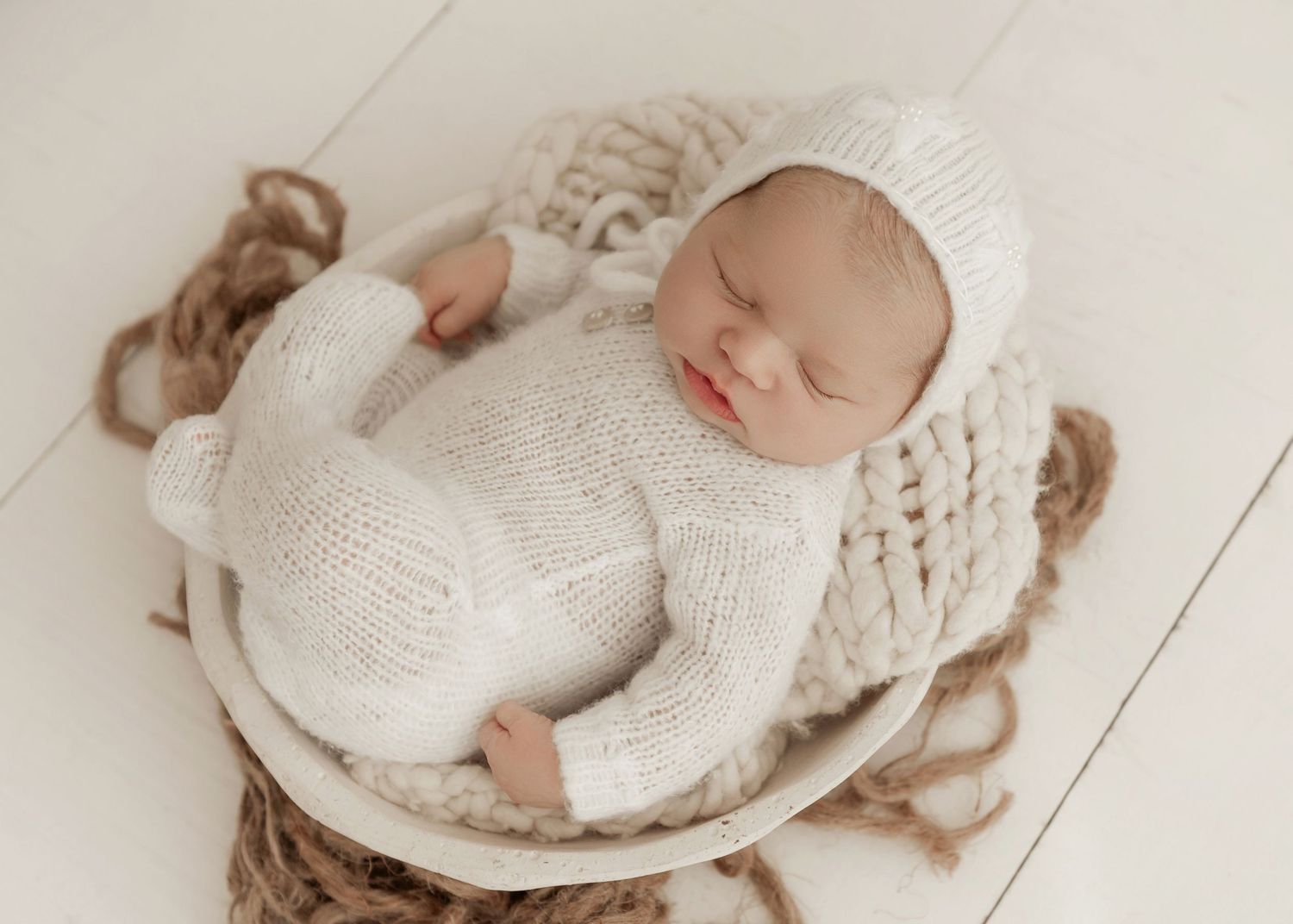 Newborn baby wrapped in white knit blanket sleeping peacefully in woven basket during professional photo session.