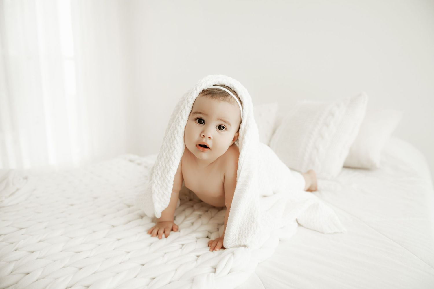 Baby wrapped in white towel crawling on soft white bedding in minimalist setting.