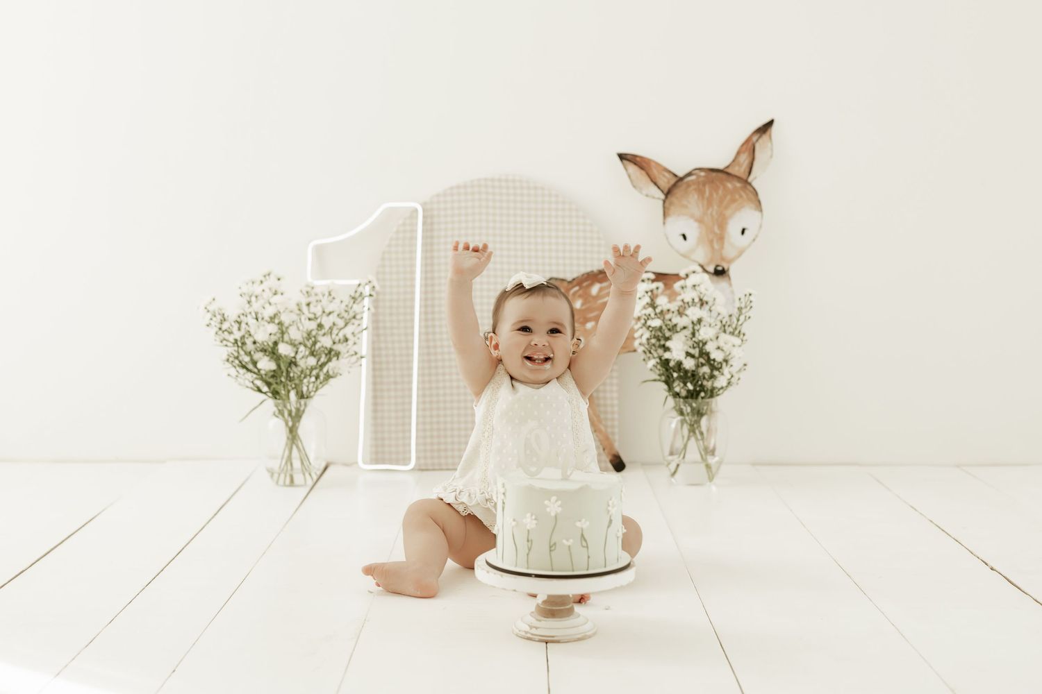 First birthday celebration setup with cake, baby's breath flowers and deer decoration on white wooden floor.