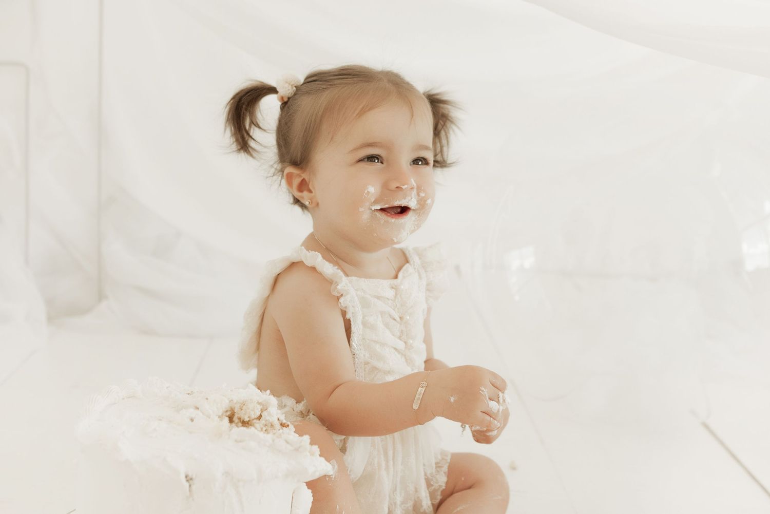 A small child in a white dress sits and smiles against a light background.