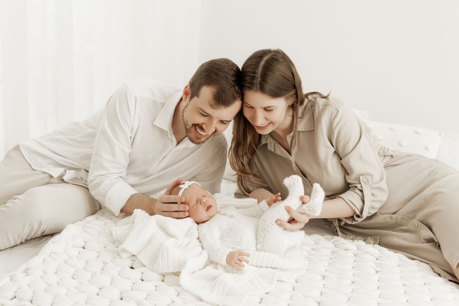 A family lies together on a white bed wearing light colored clothing while sharing tender moments.