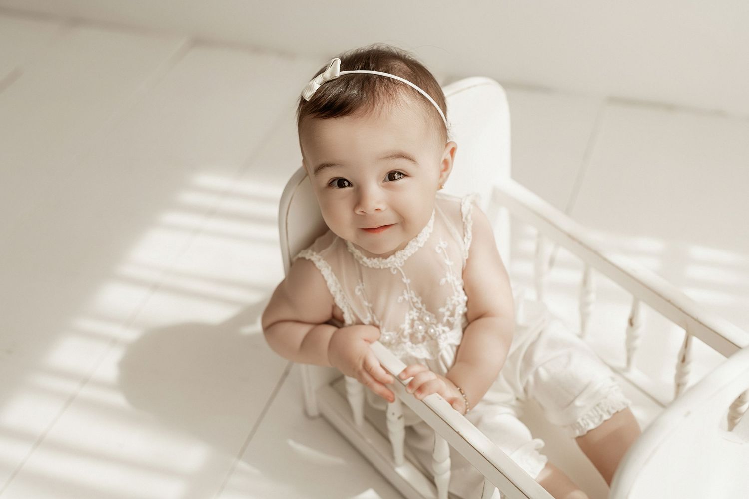 A baby in a white lace dress sits in soft window light creating a dreamy portrait series.
