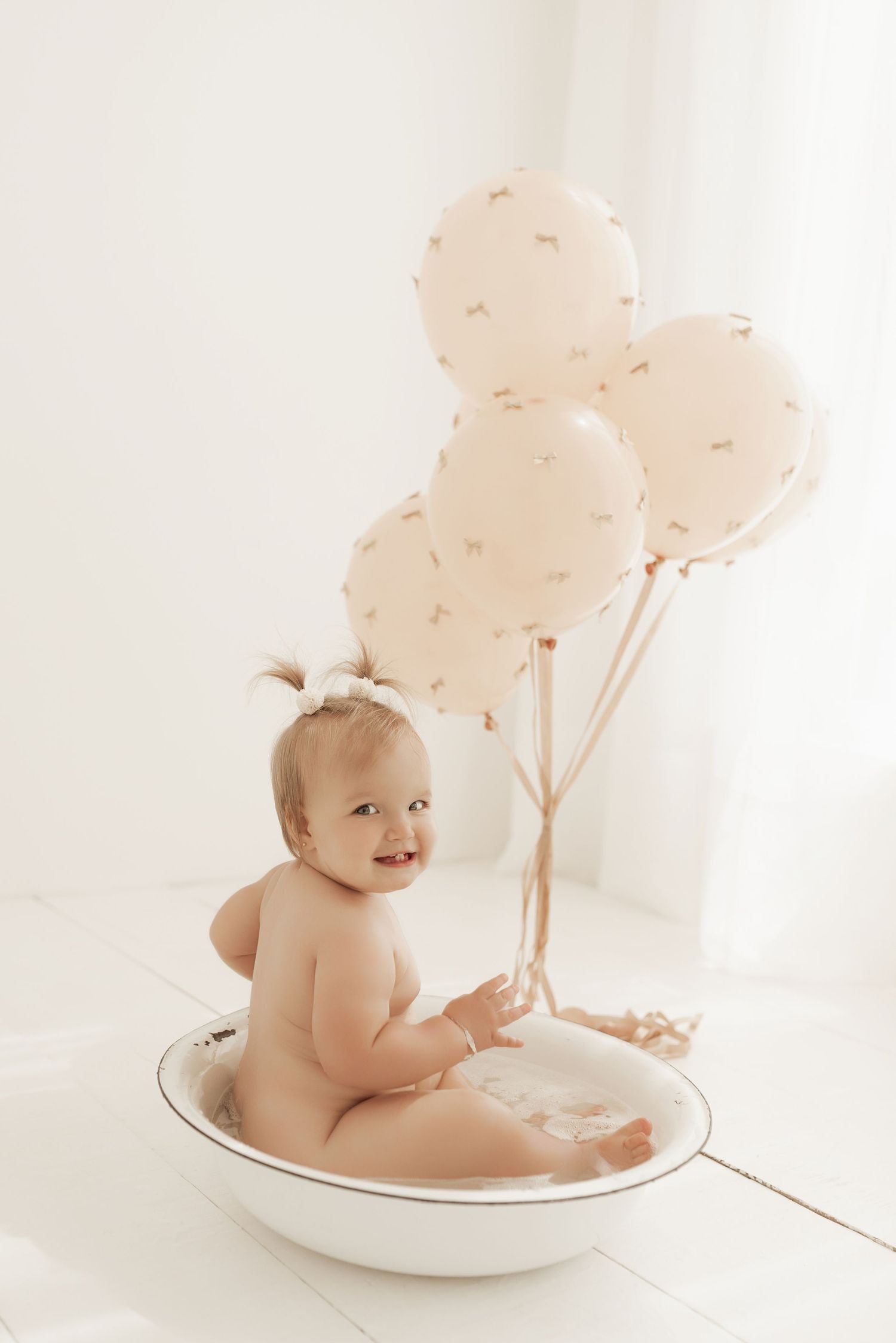 A baby sits in a white bowl against a light backdrop with soft cotton ball decorations in a minimalist photo setting.