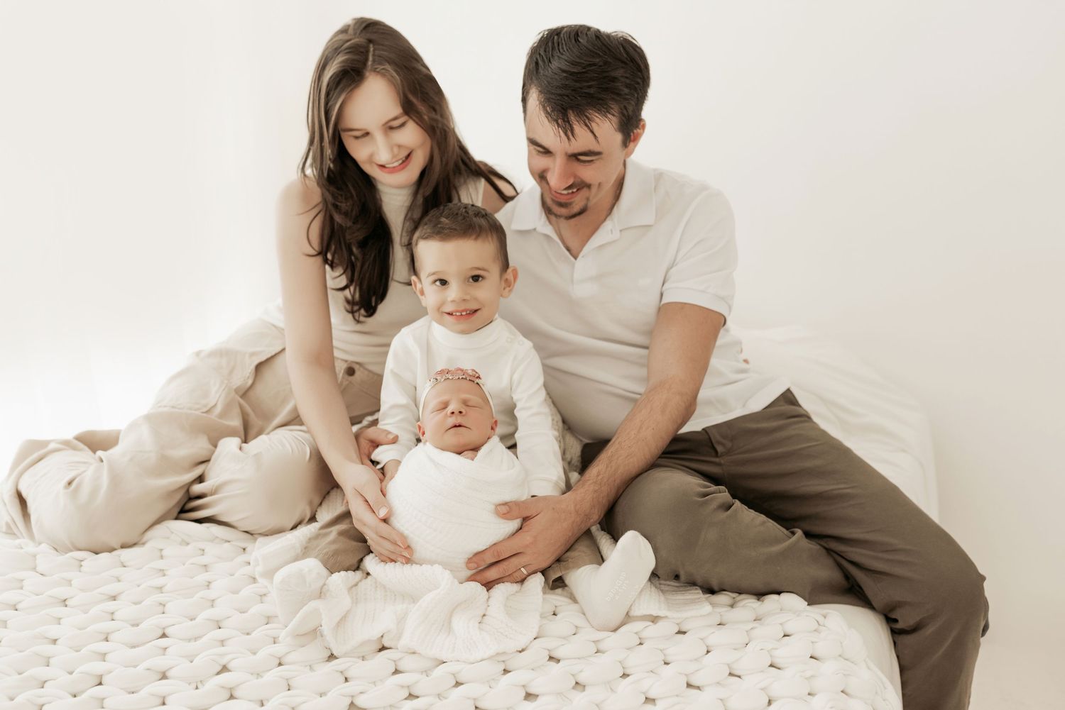 Family in neutral colored clothing sits together on textured white blanket during intimate portrait session.