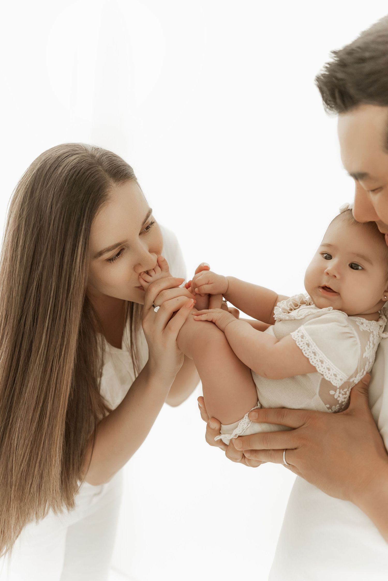 Intimate family moment showing parents in white clothing playing with their baby wearing a delicate lace dress.