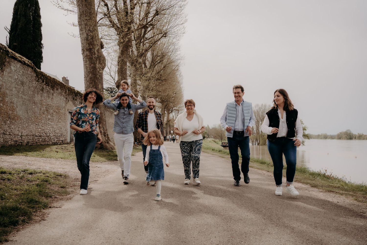 Family group walking together on tree-lined path by stone wall.