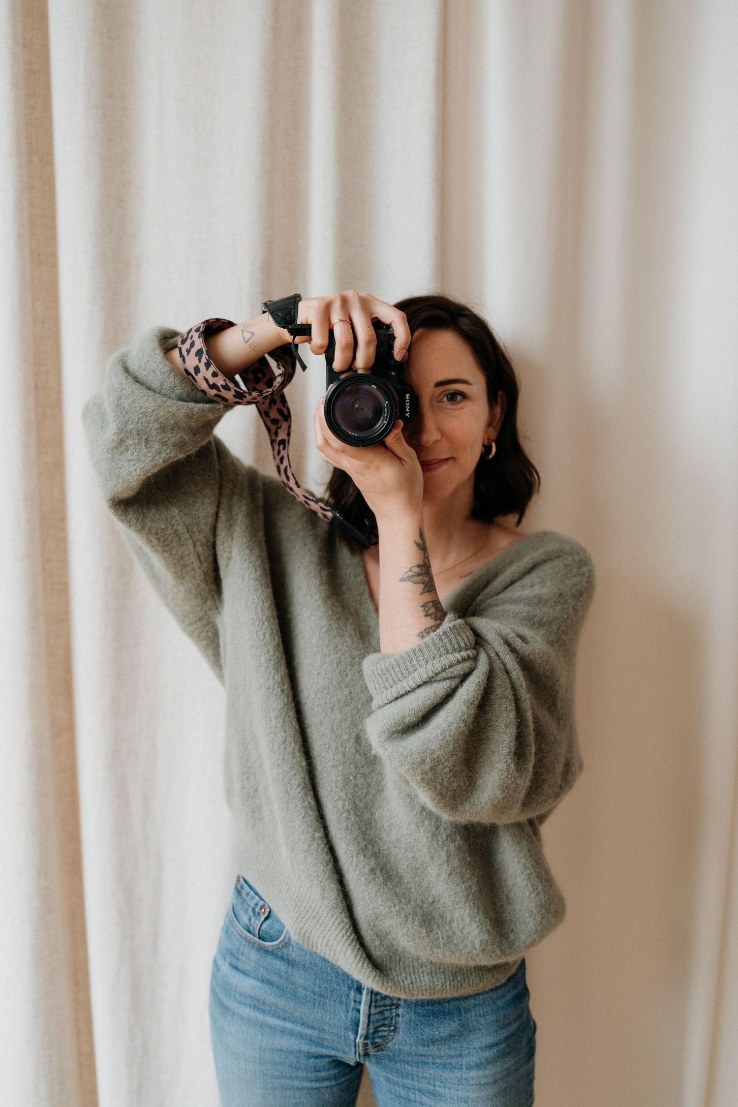 A photographer in a gray sweater takes a photo against white curtains.