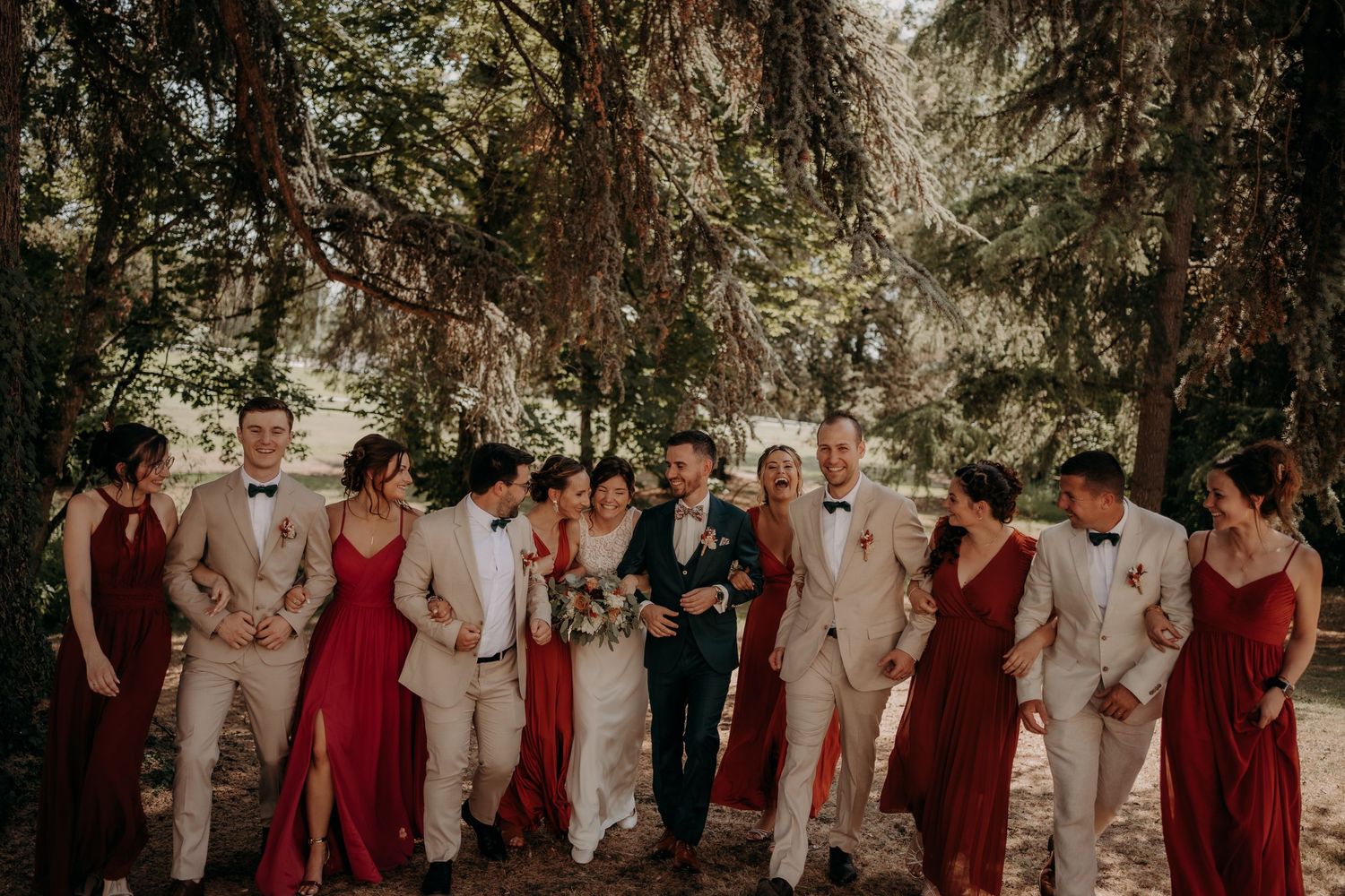 A wedding party poses together outdoors wearing burgundy dresses and beige suits among tall evergreen trees.