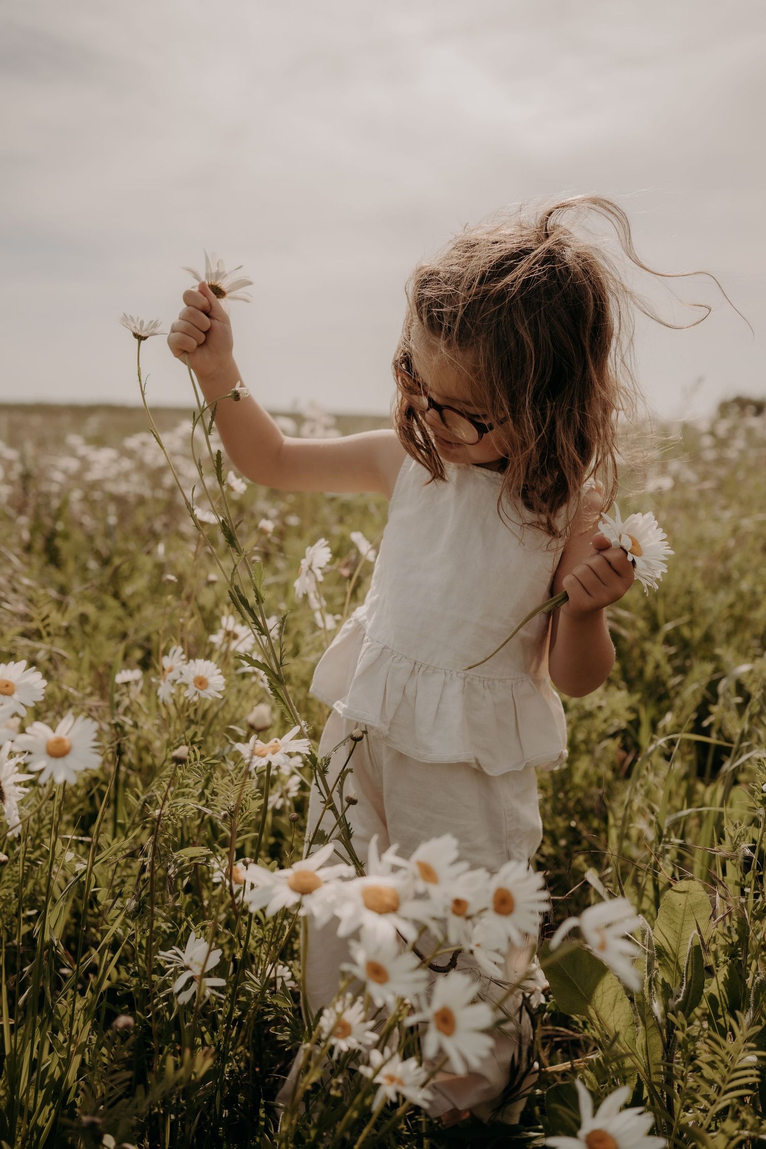 Young child in white dress playing among wild daisies in summer meadow.