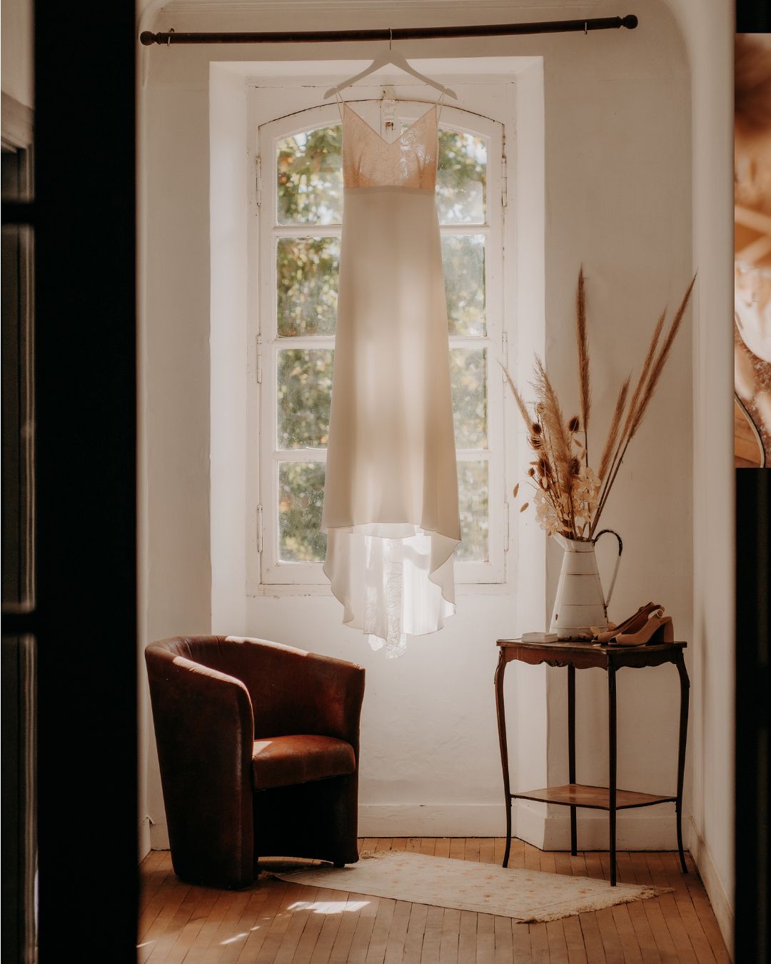 A white wedding dress hangs elegantly in front of an arched window, with a brown leather chair and side table nearby.