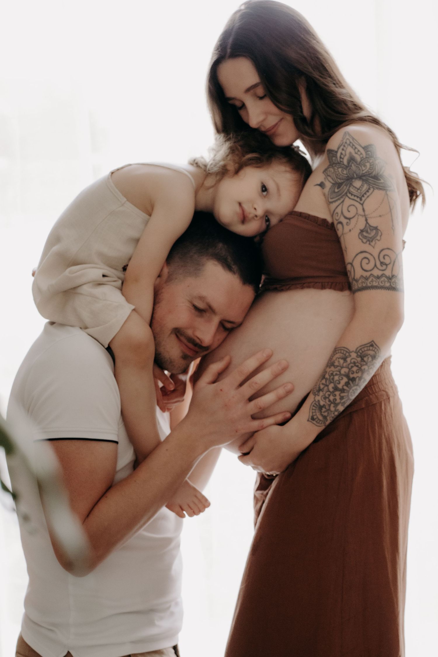 Emotional maternity session with couple in white and brown clothing against bright background.
