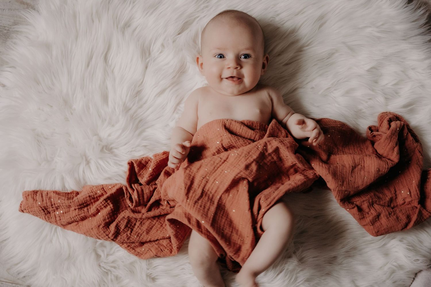 Baby lying on white fur rug with terracotta colored fabric.