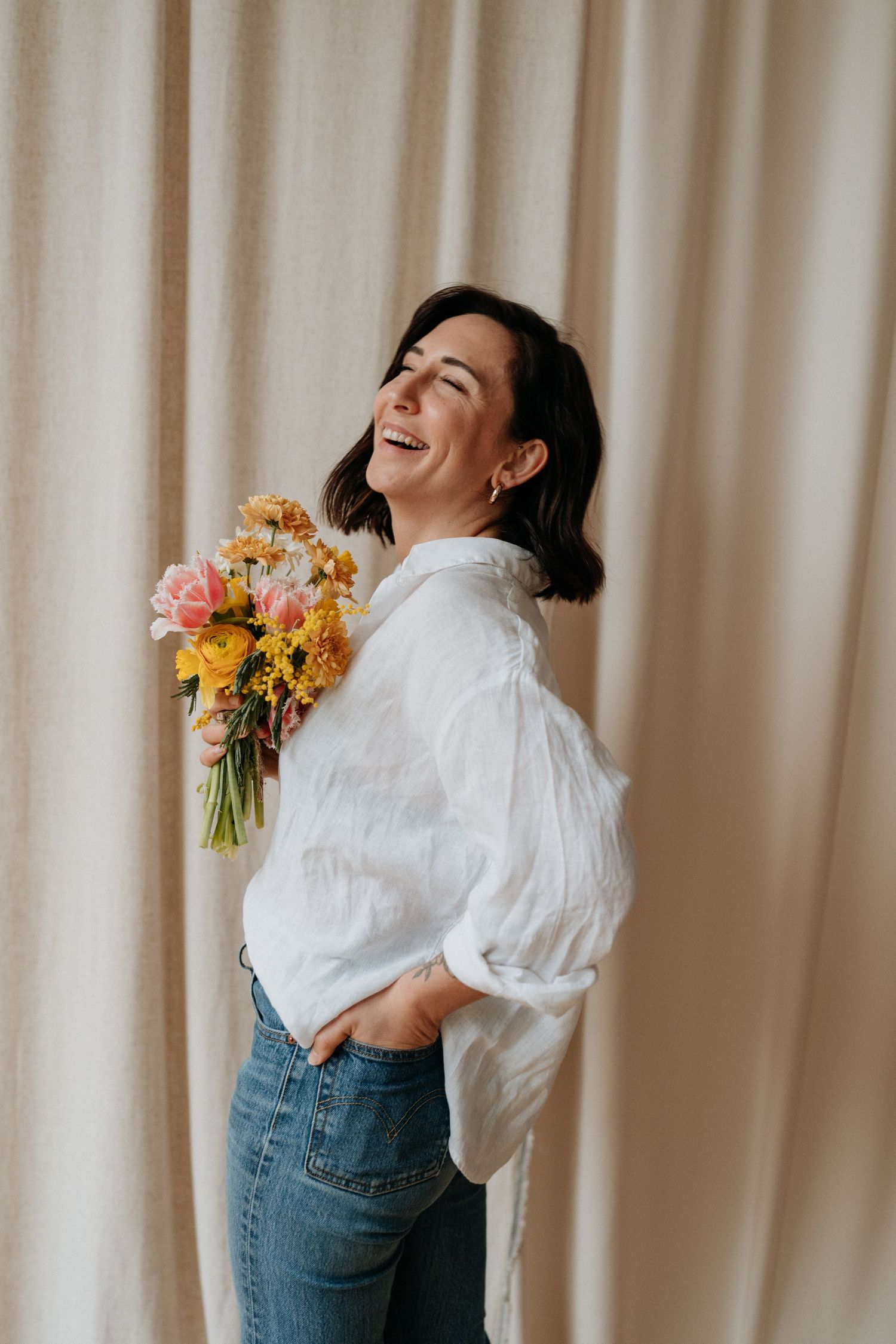 Woman in white blouse and jeans holding colorful flowers against a light curtain background.