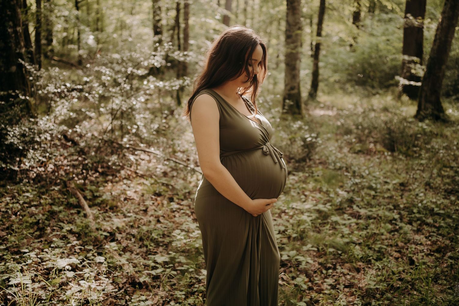 Expectant mother in brown dress standing in sunlit forest setting.