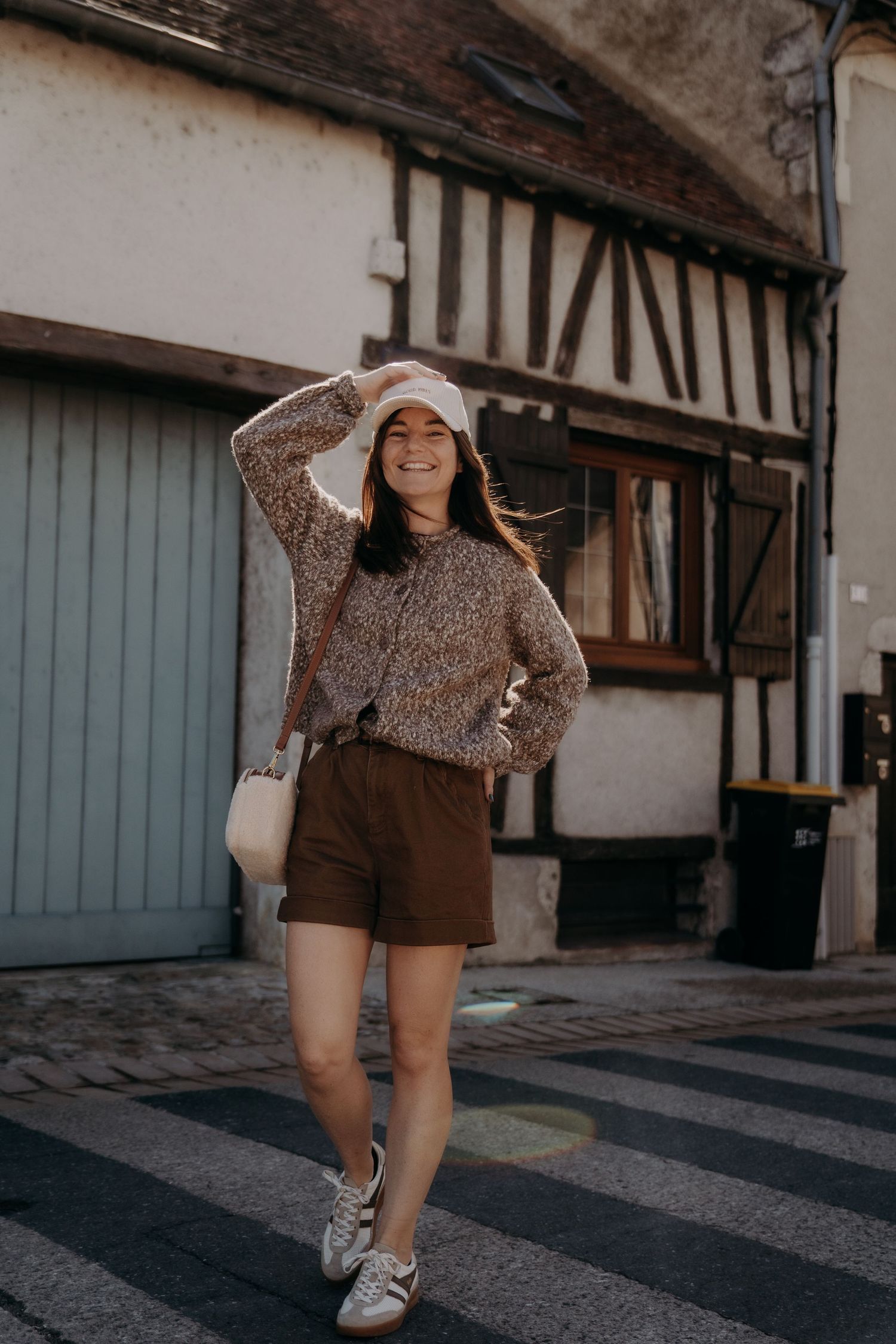 Person in brown shorts and sparkly sweater posing playfully on cobblestone street.