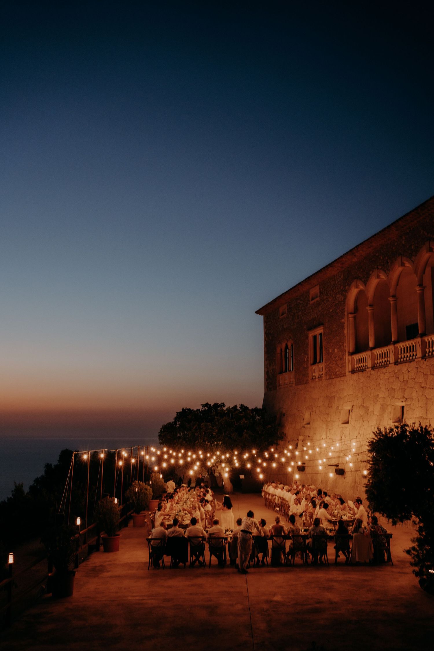 Outdoor wedding reception with string lights at sunset against Mediterranean villa.