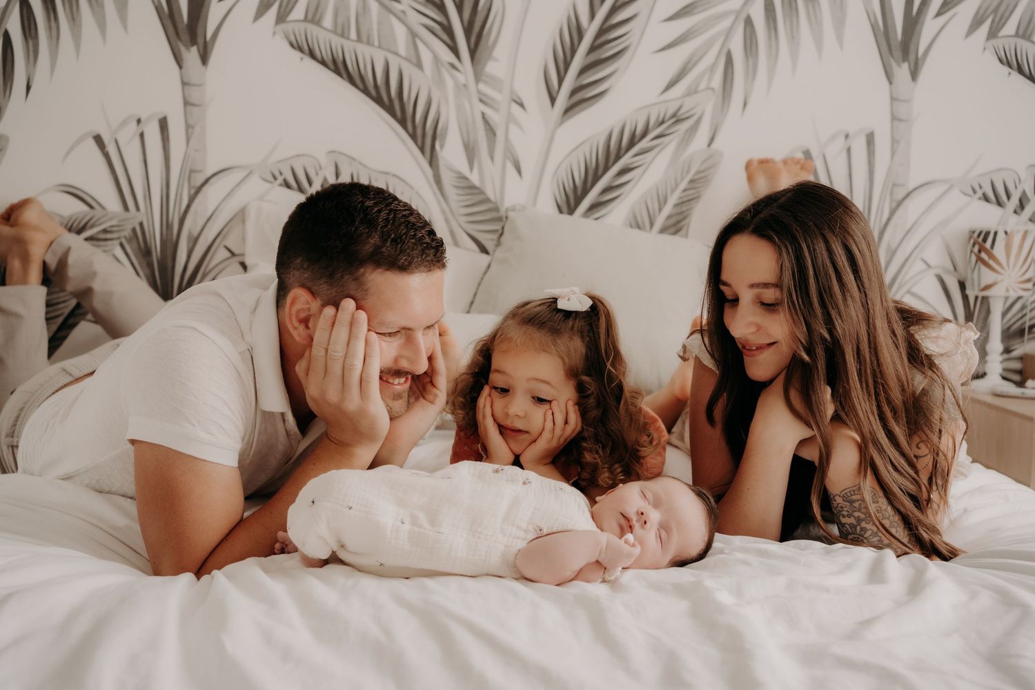 Family cuddles on bed with tropical leaf wallpaper backdrop.
