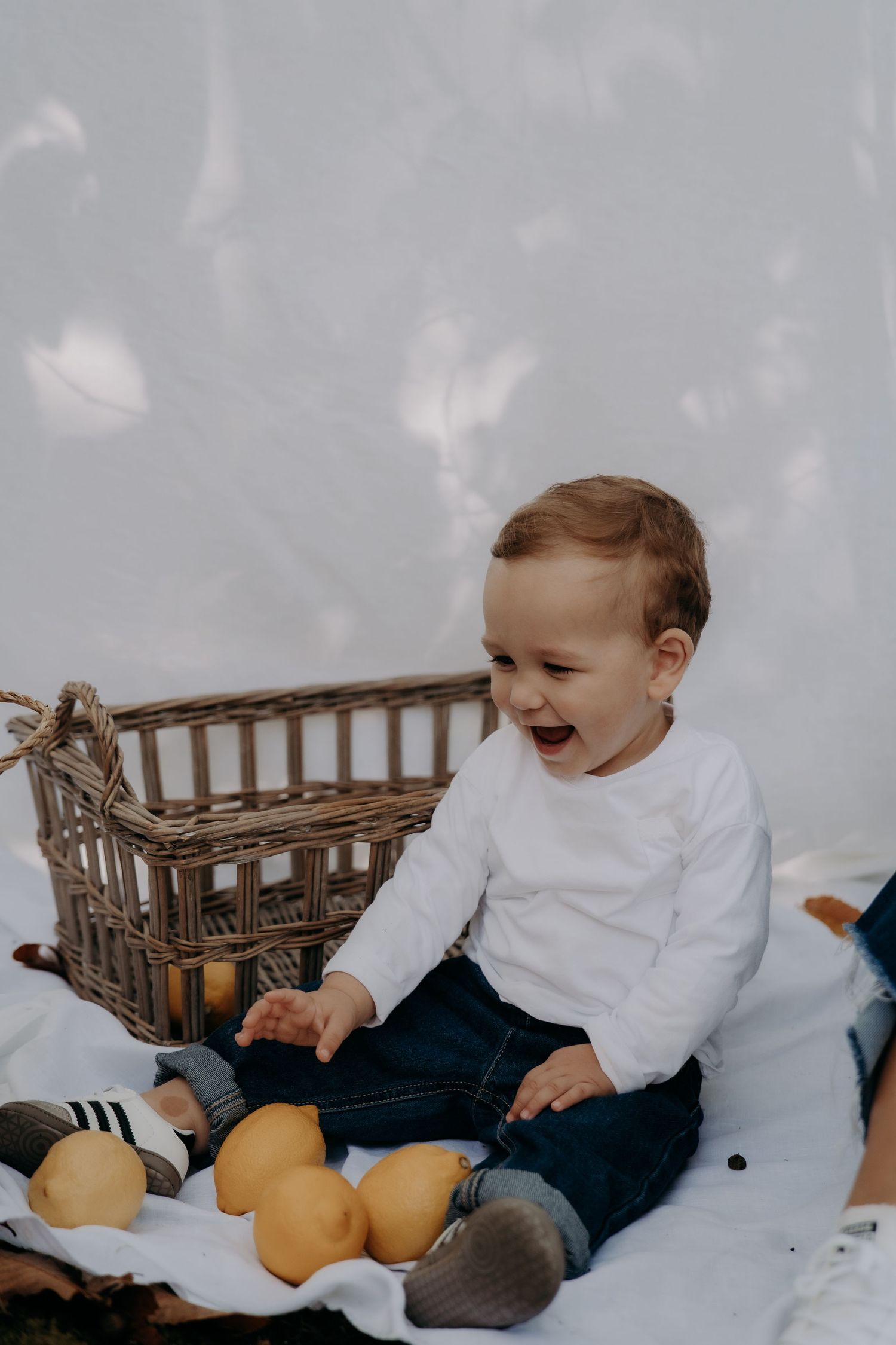 Laughing toddler in white shirt sits on blanket next to wicker basket with citrus fruits.