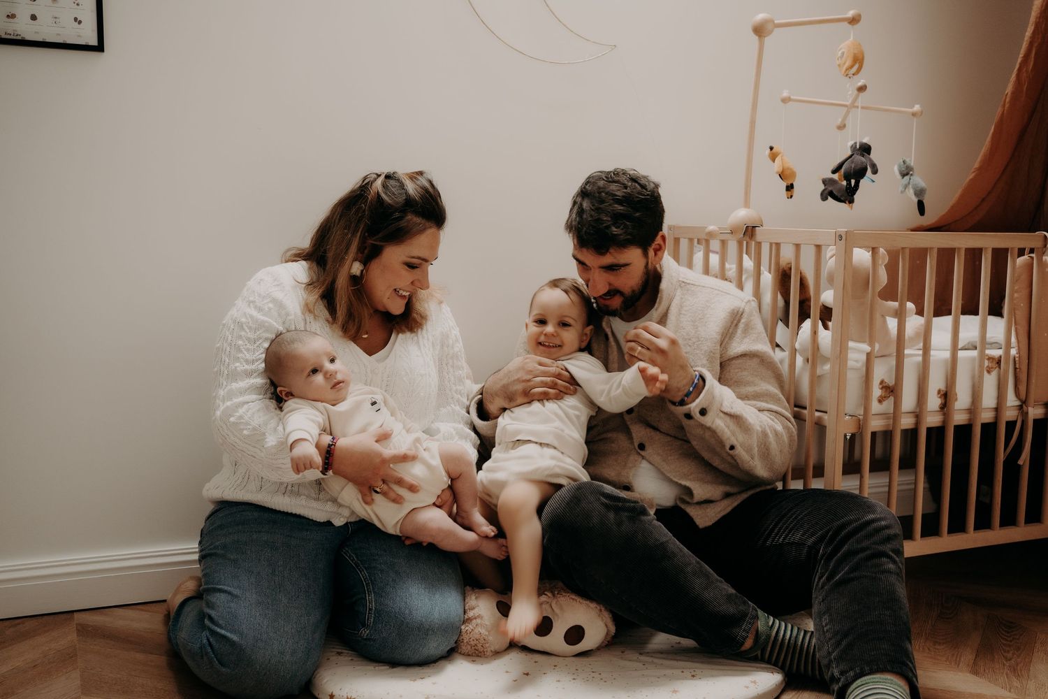 Family sitting together on floor of nursery with wooden crib in background.