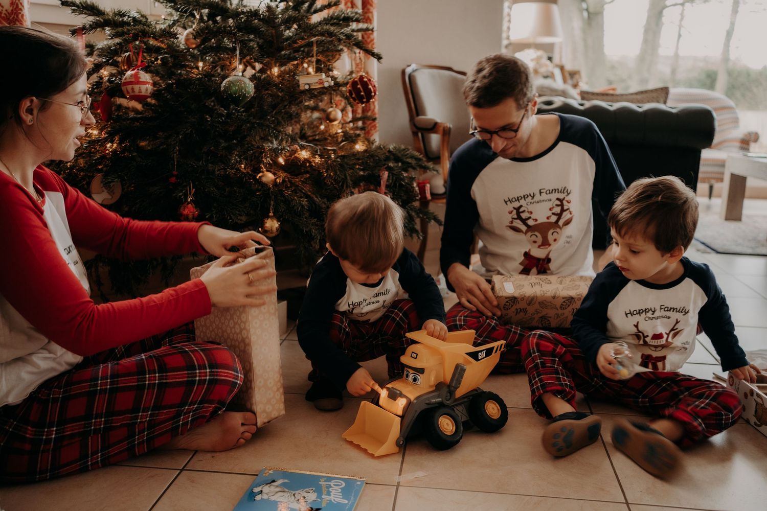 Family in matching holiday pajamas playing with toys by Christmas tree.