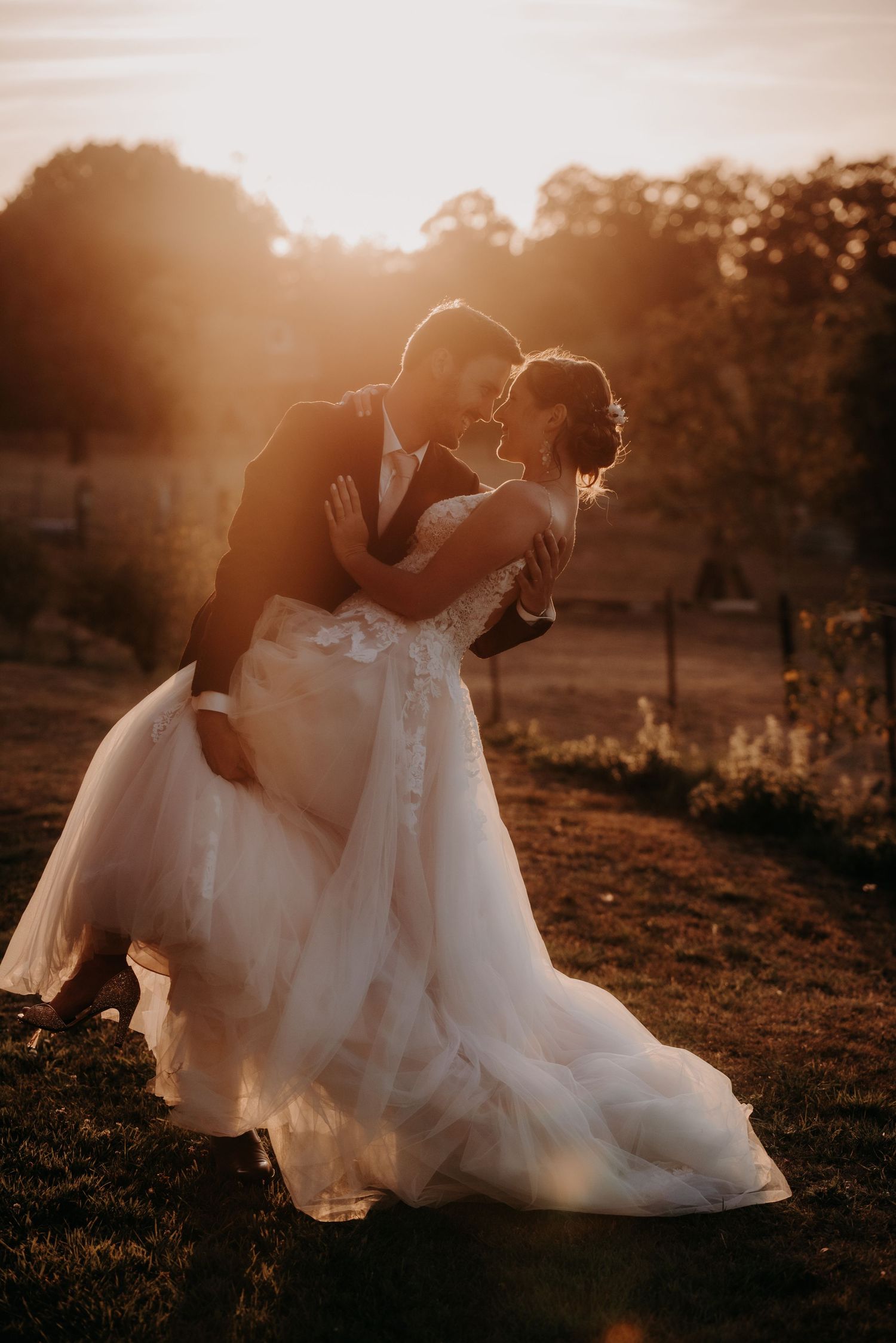 Newlyweds embrace on a grassy hill during golden hour with dramatic sunlight.