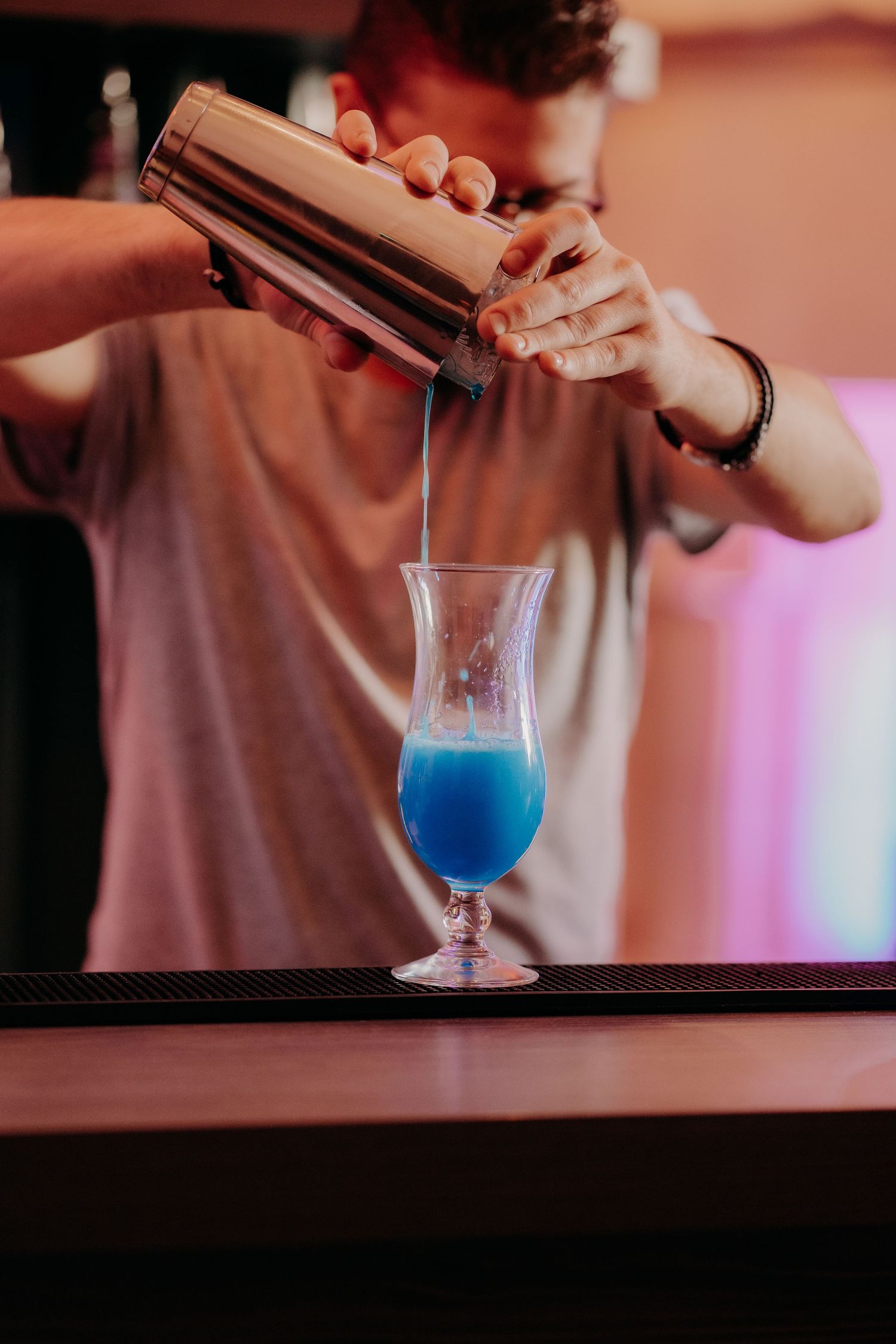 Hands pouring bright blue cocktail into a glass with pink ambient lighting.