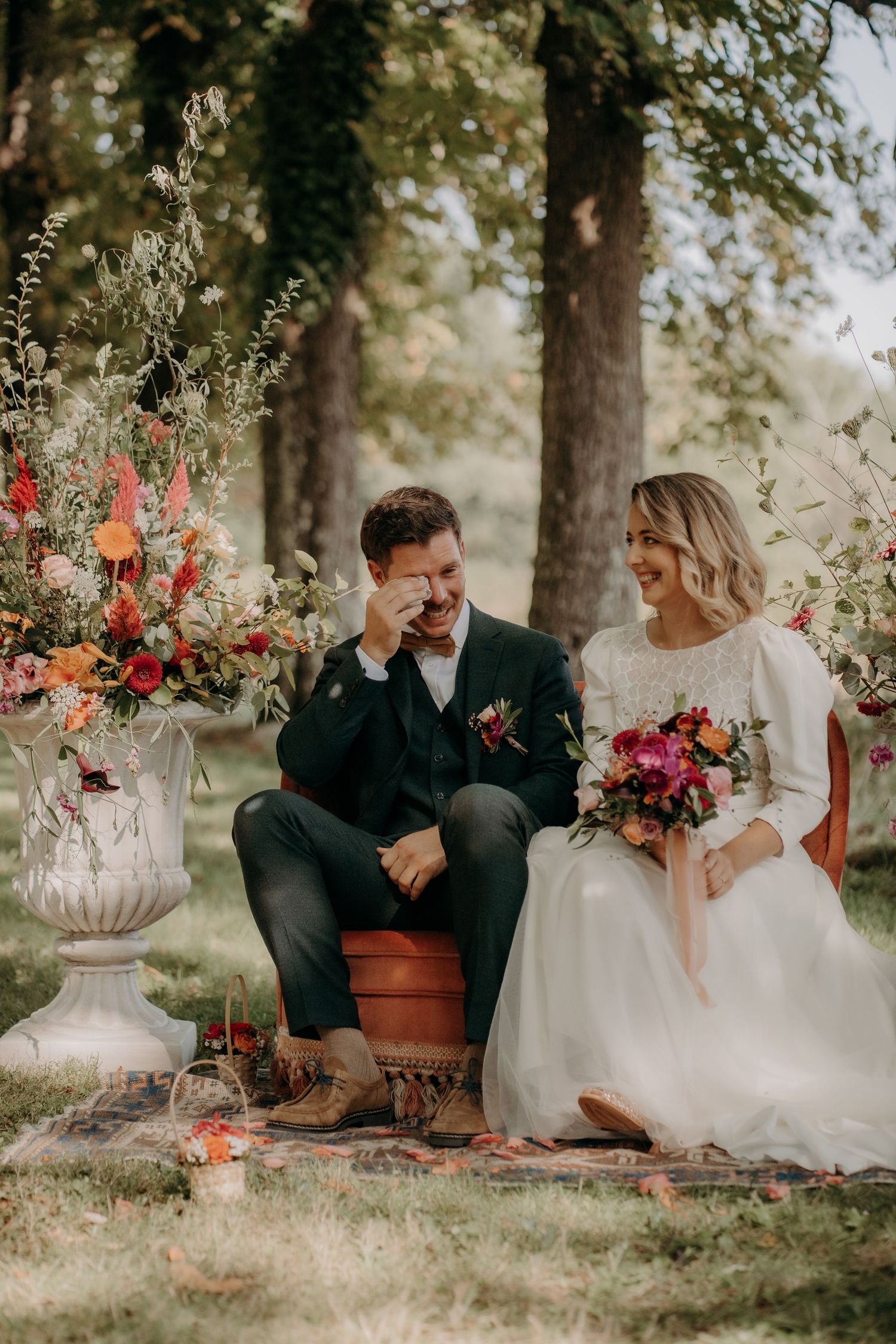 Newlyweds share intimate moments on rustic bench surrounded by vibrant floral arrangements.