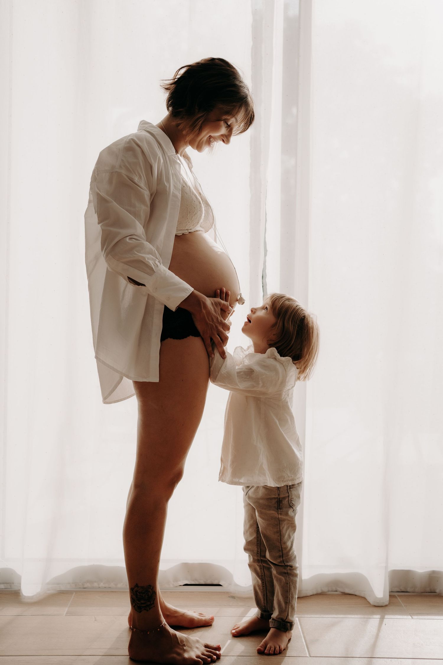 Touching maternity moment by window with white curtains and natural light.