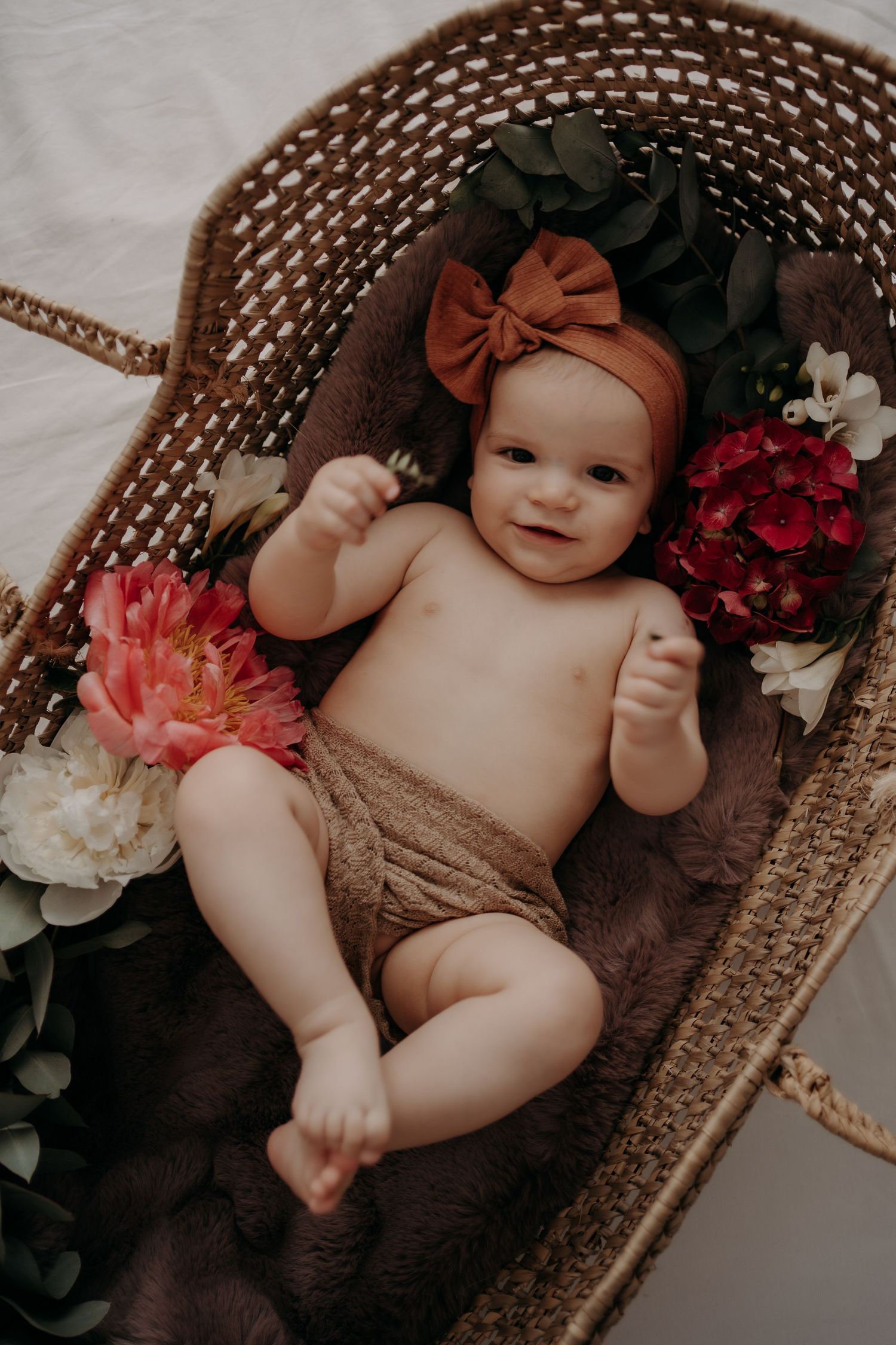 Baby lying in wicker basket surrounded by flowers and wearing orange bow headband.